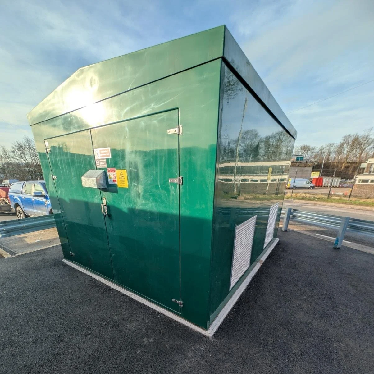 Green electrical transformer box on asphalt parking lot, with a silver warning sign, locks, and safety labels, parked beside a metal guardrail.