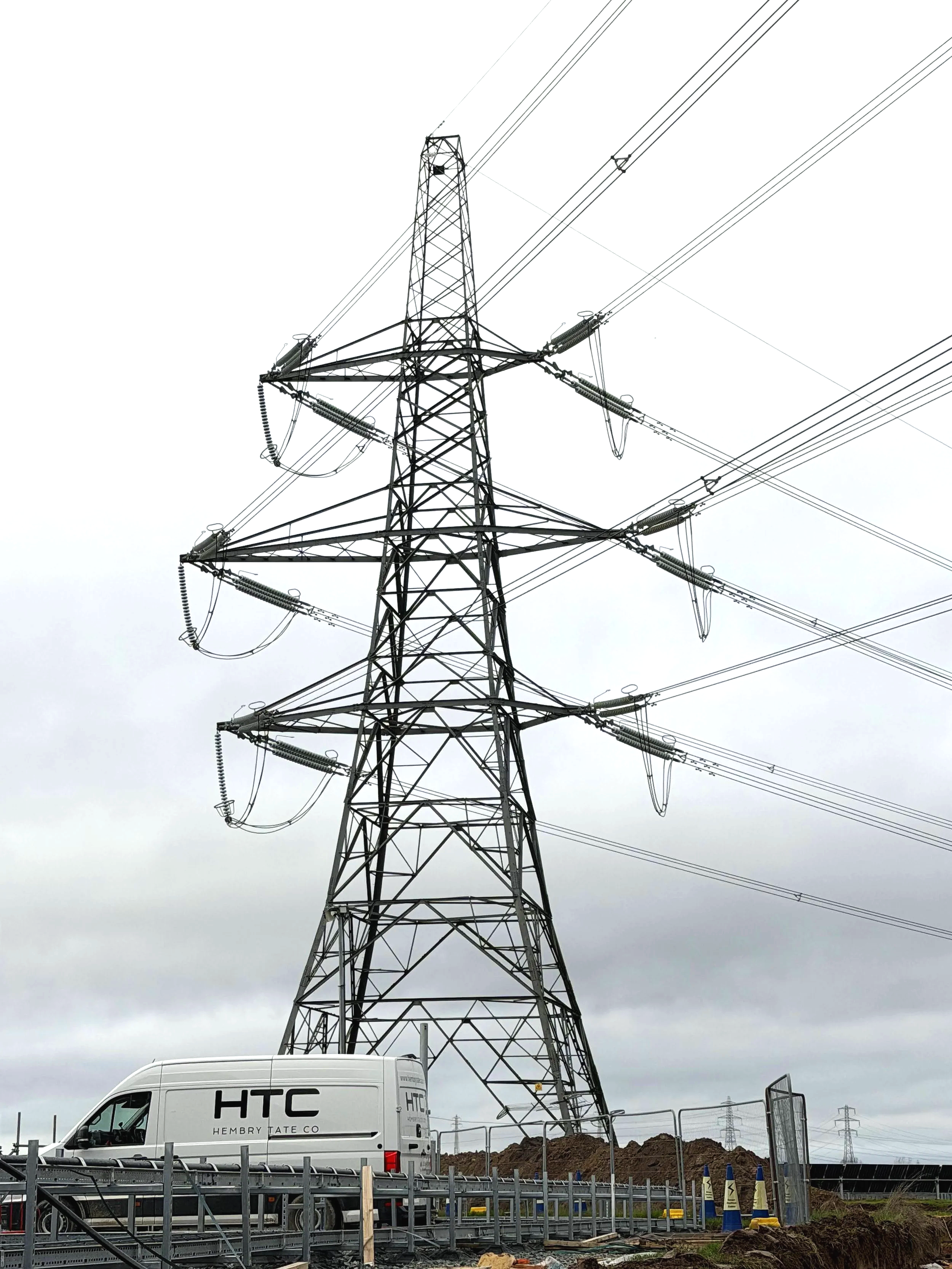 A tall electrical transmission tower with multiple power lines extending from it, set against a cloudy sky, near a construction site with a white van labeled HTC Hembry Tate Co.
