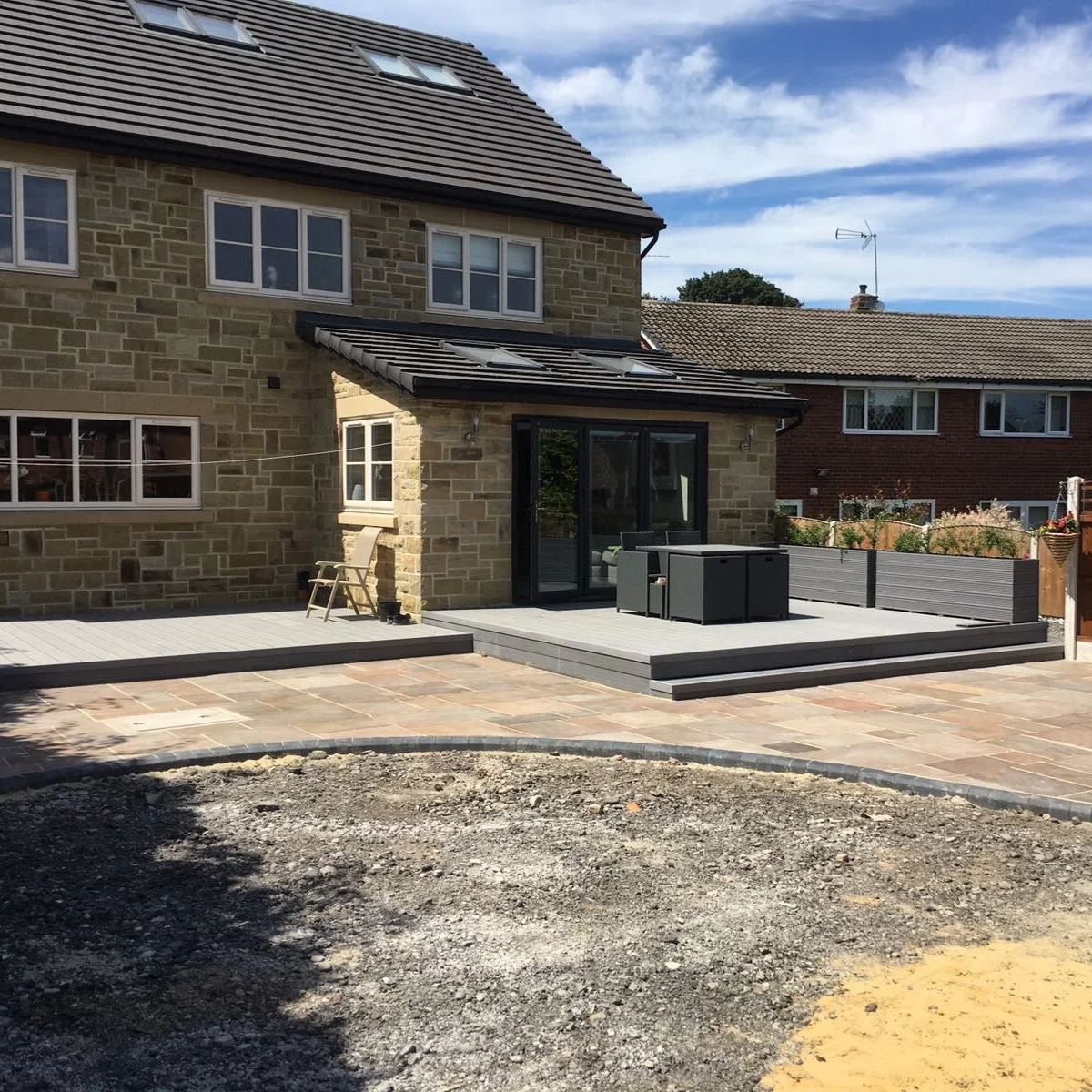 View of a newly renovated backyard patio and deck area attached to a multi-story brick and stone house. The patio has stone paving, and the deck has outdoor furniture, with a small chair near the house.