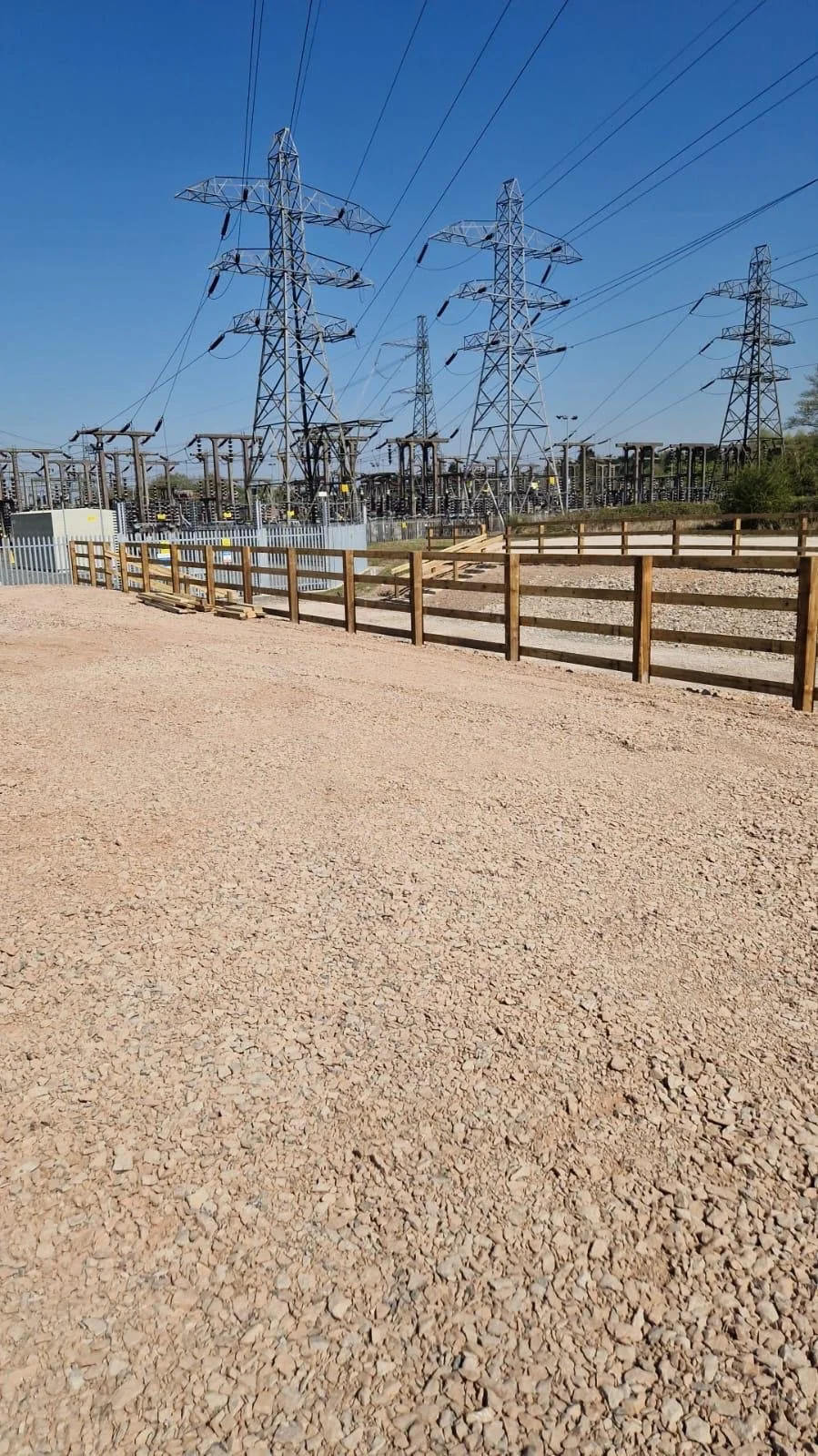 View of high voltage power lines and electrical towers in an electrical substation, with a dirt ground and a wooden fence, under a clear blue sky.