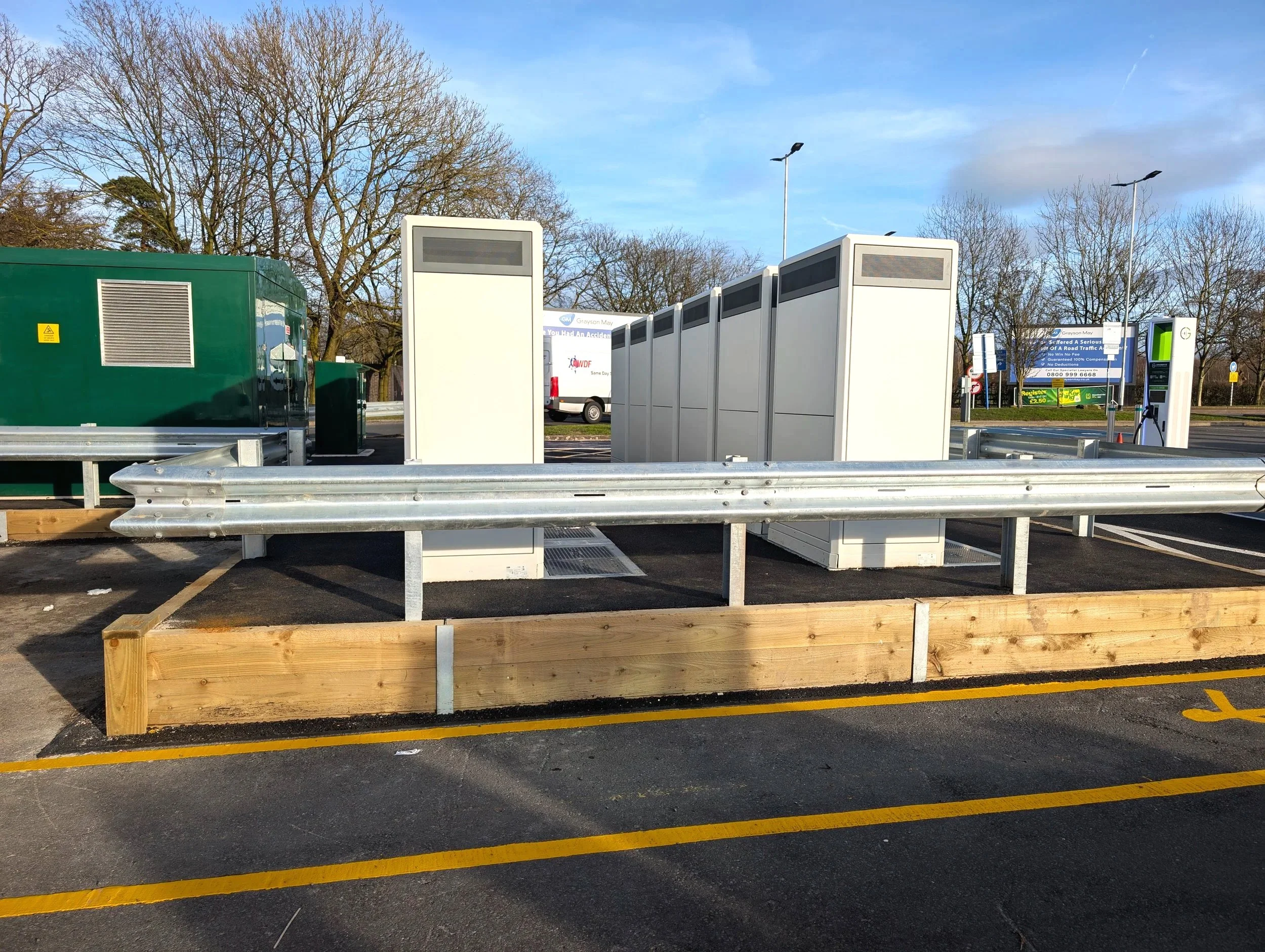 Electric vehicle charging station with multiple charging units, a green generator, and a parking lot with safety barriers and trees in the background.