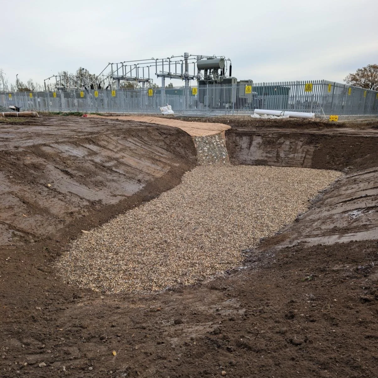 Construction site with a large excavation area filled with small gravel, surrounded by dirt and a metal fence.