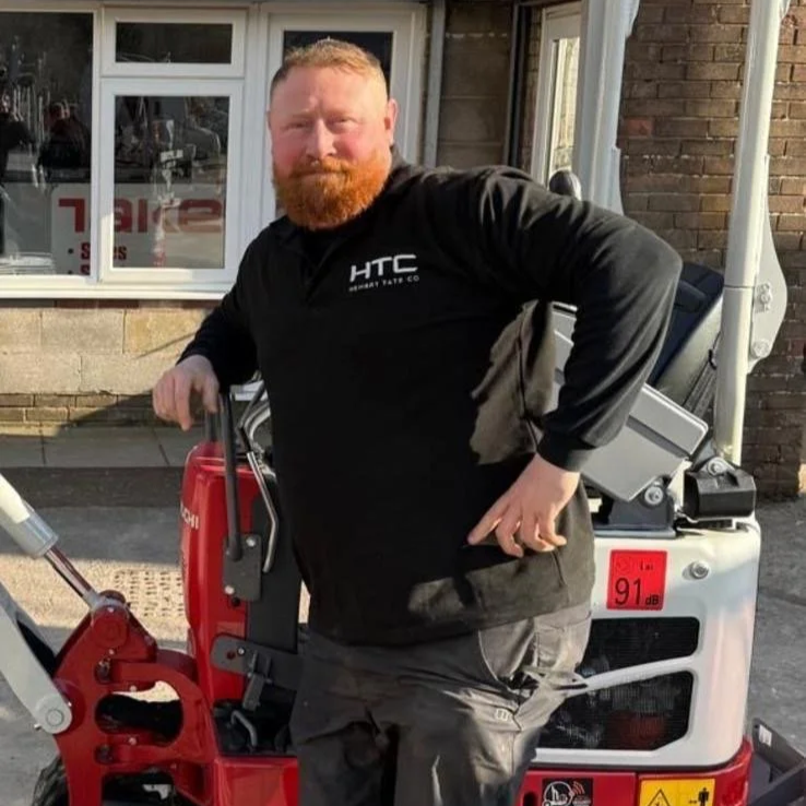 A man with a beard in a black HTC jacket standing outdoors next to a red and white piece of equipment.