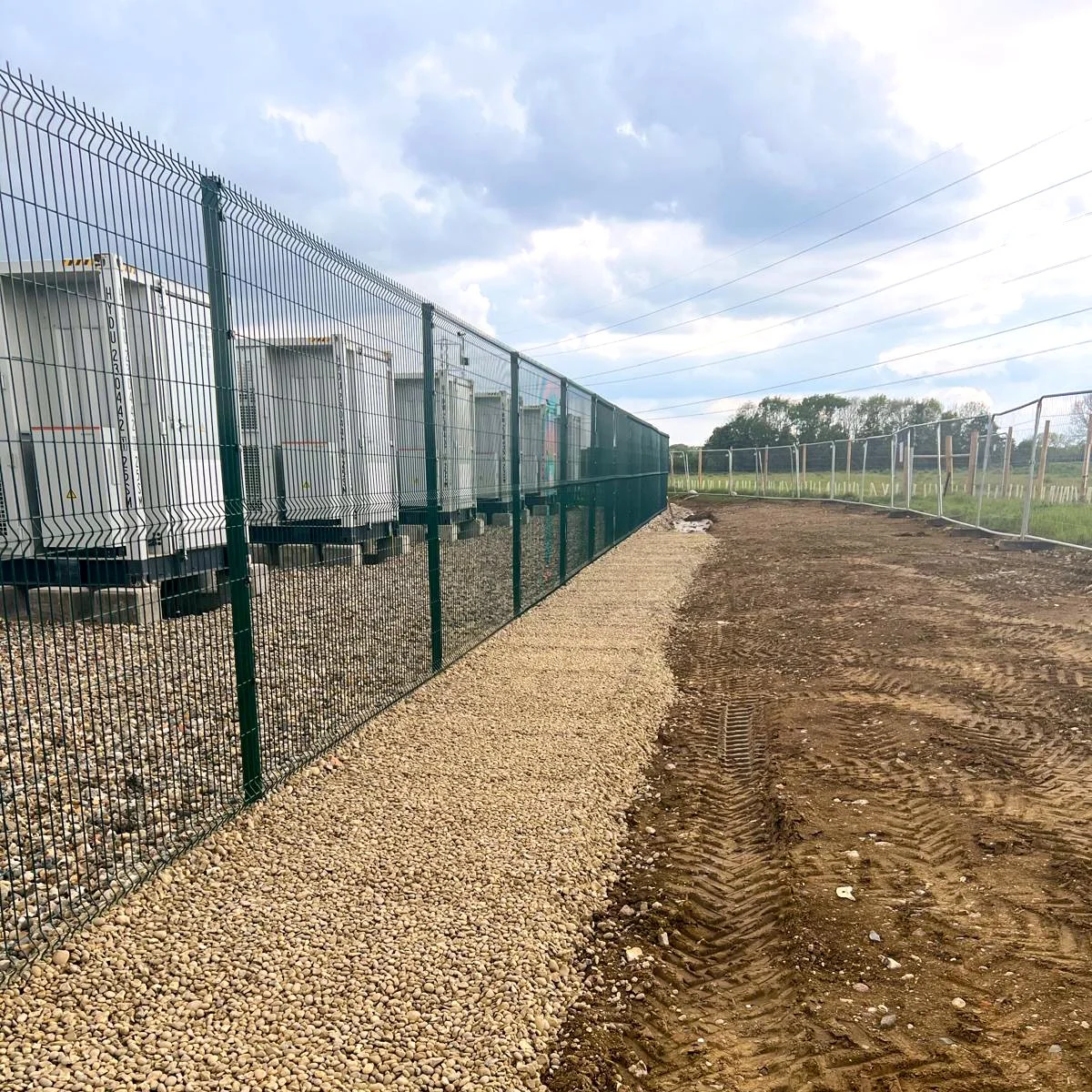 Construction site with a new gravel pathway beside a green fence, enclosing electrical transformers, under a cloudy sky.