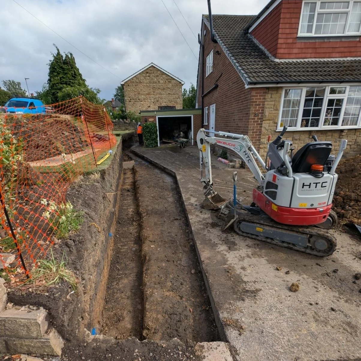 Construction site with a mini excavator, orange safety fencing on the left, and a partially dug trench beside a house with brick walls and a checkered roof. Worker in high visibility vest in the background near a garage.
