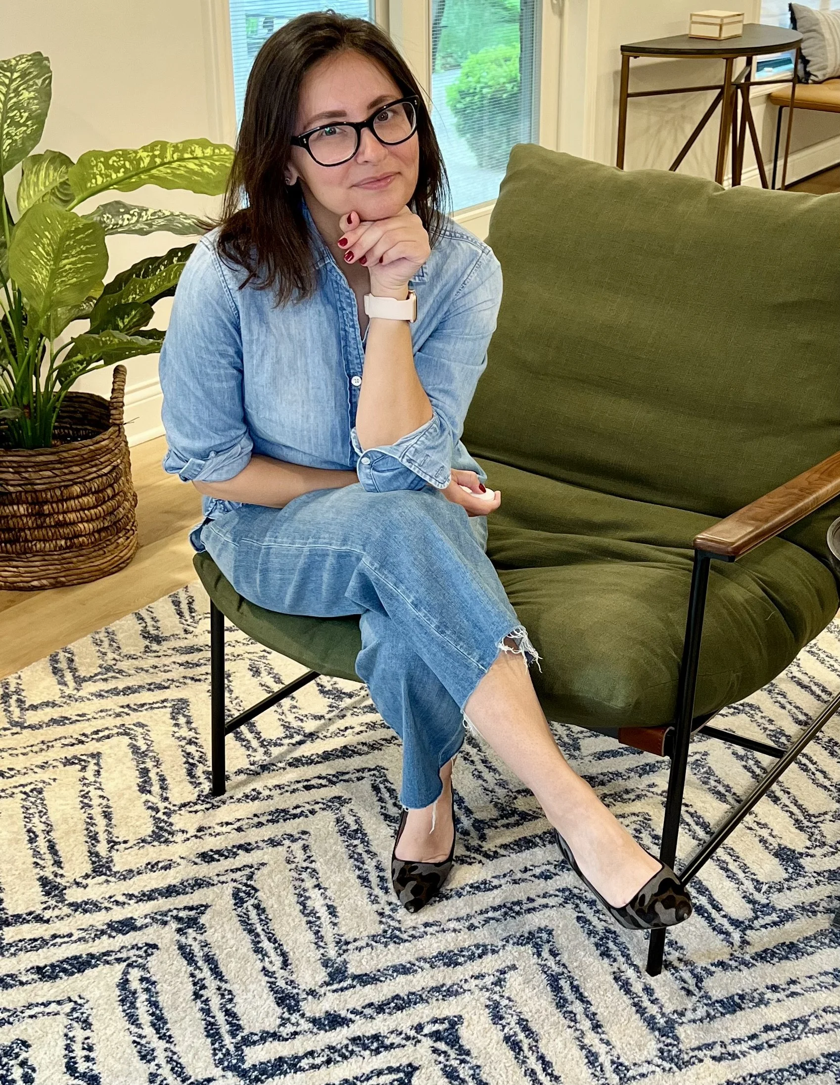 A woman with dark hair, glasses, and red nails sitting on a green chair with her legs crossed, wearing a light blue denim shirt, ripped jeans, and leopard print shoes, next to a large potted plant and a window with a view of greenery.