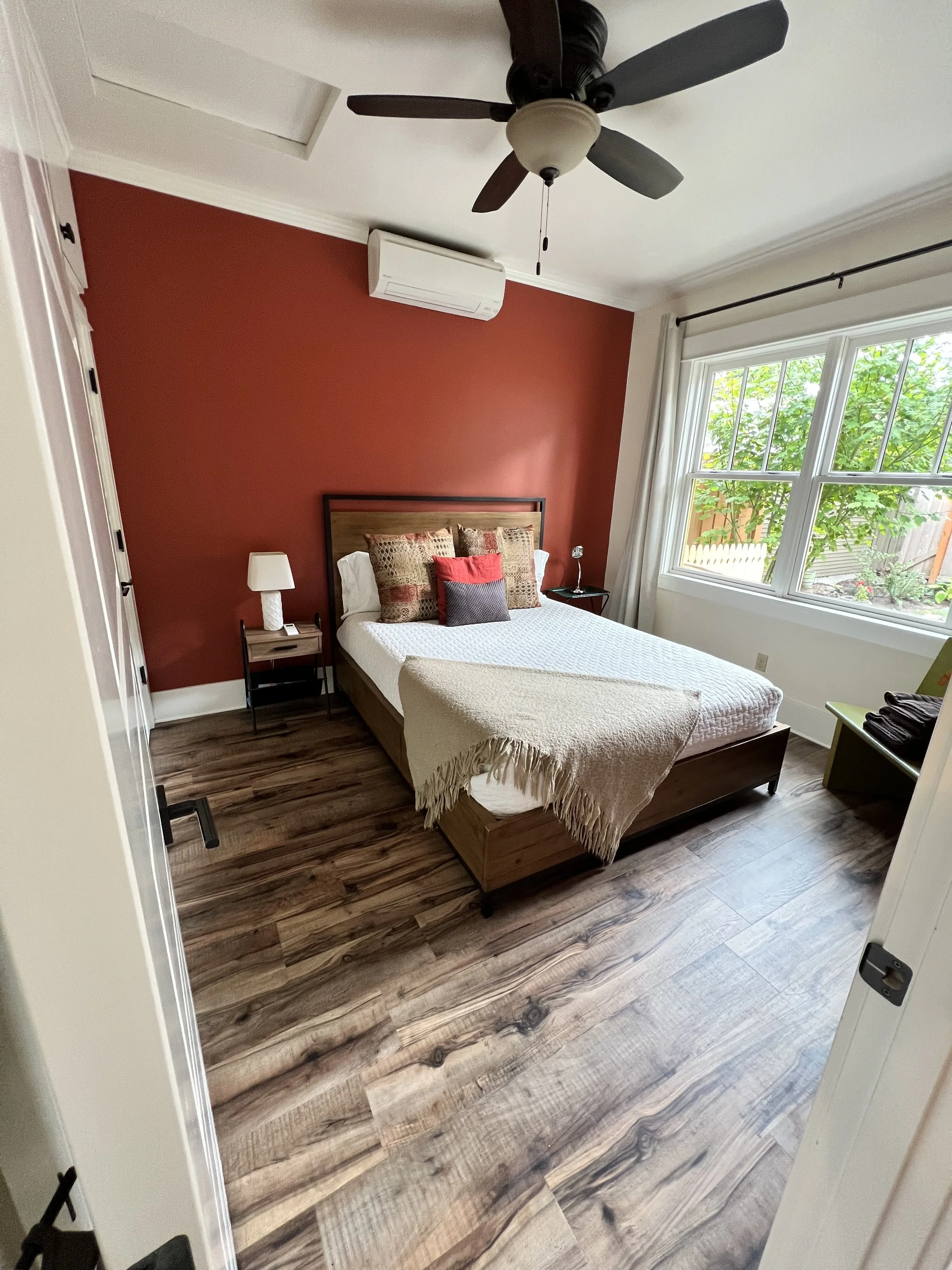 A bedroom with a large window, a bed with a wooden headboard, and a red accent wall. There are nightstands with lamps on either side of the bed, a ceiling fan with dark blades, and hardwood flooring.