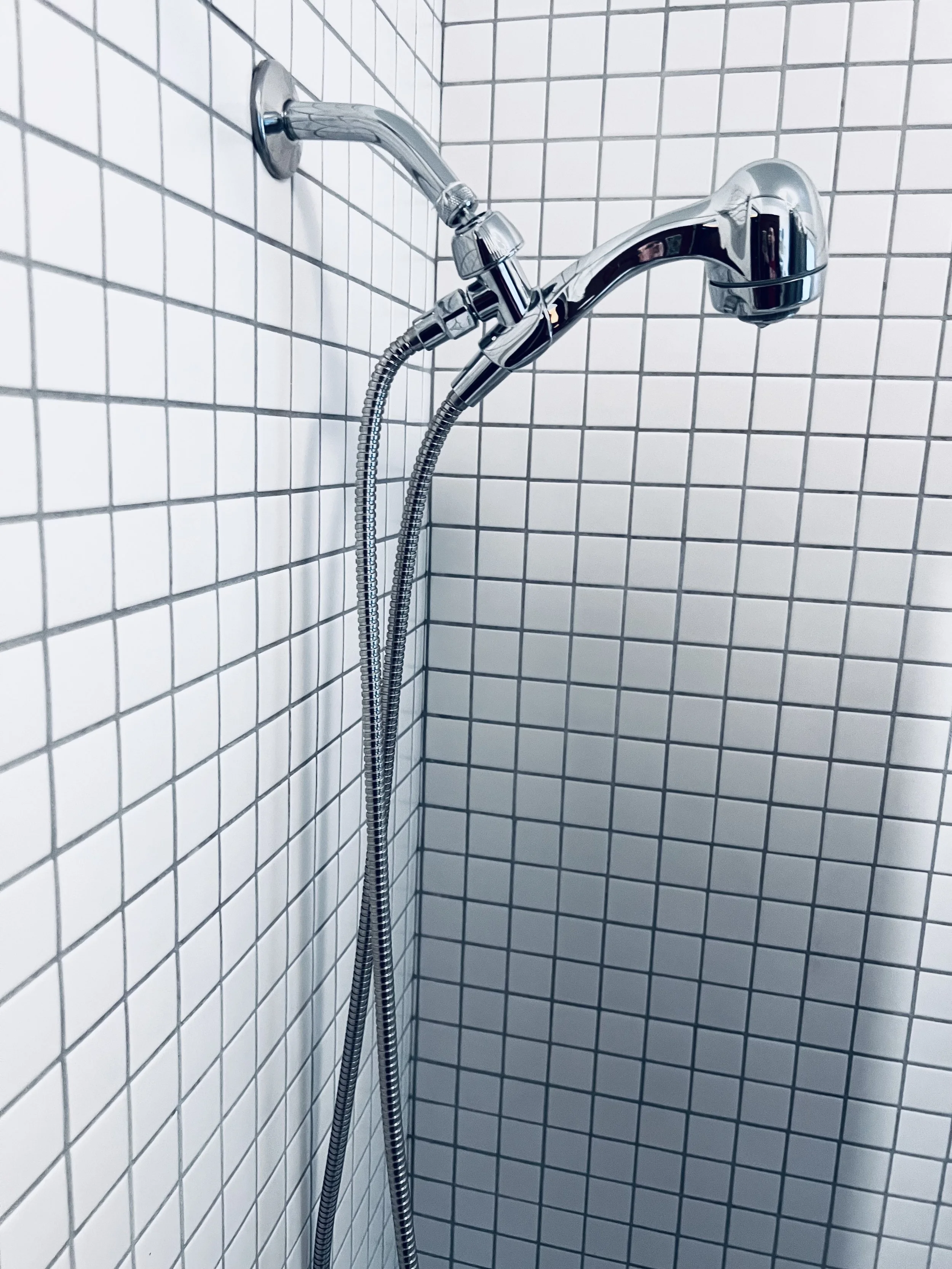 Close-up of a chrome showerhead and handheld shower wand attached to a tiled bathroom wall with white square tiles.