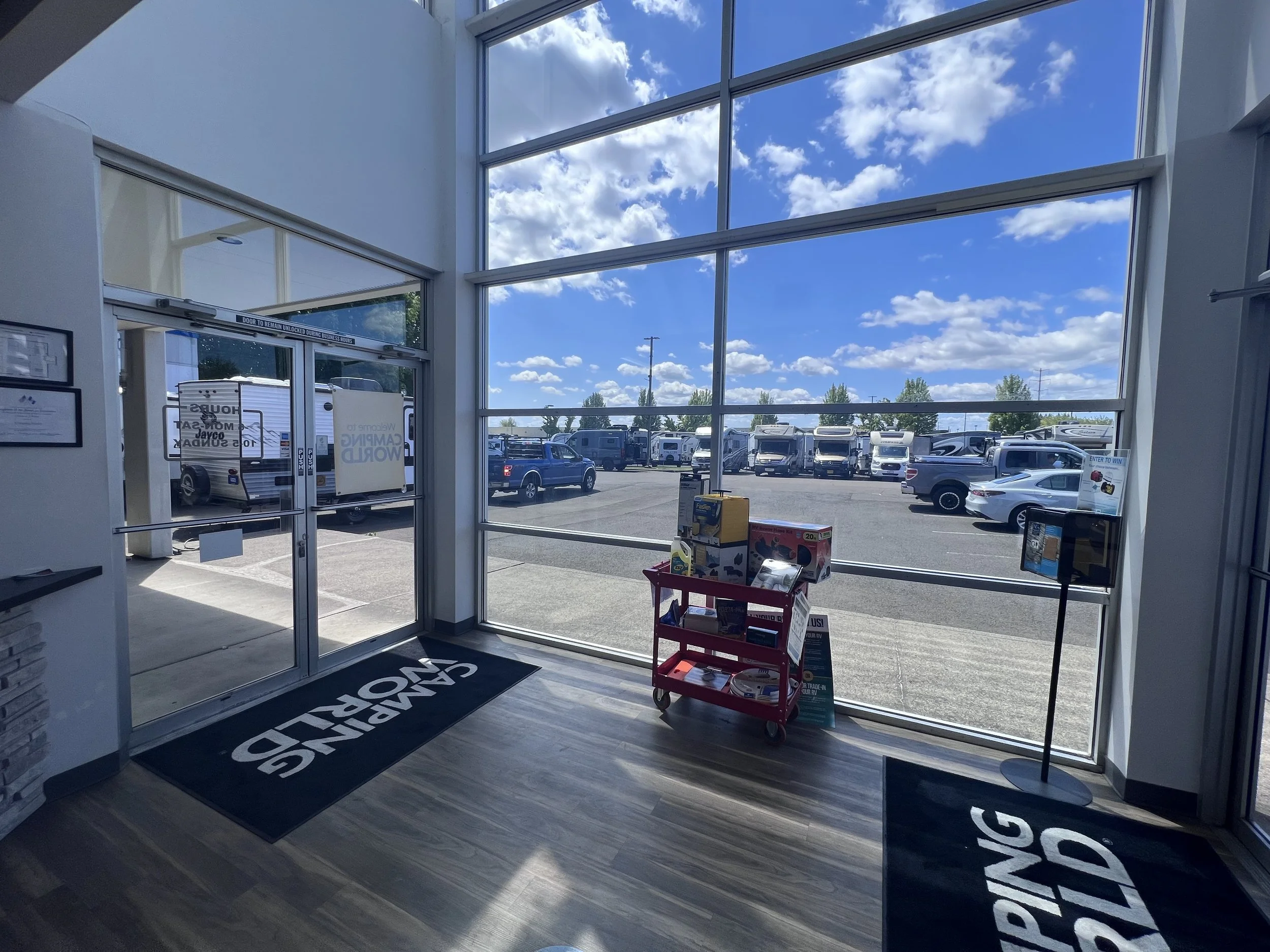 Interior view of a showroom with large glass windows showing outdoor parking lot, clear sky with clouds, and RVs parked outside. Inside features a red cart with boxes and a standing display.
