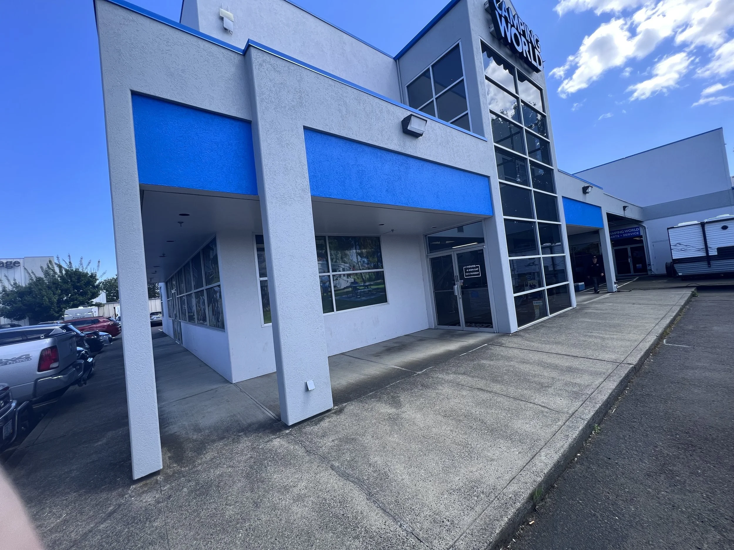 The storefront of a retail or electronics store with a blue and white exterior, large glass windows, and a sign that reads 'Gaming World' at the top. There is a parking lot with several cars in front and a clear blue sky with some clouds overhead.