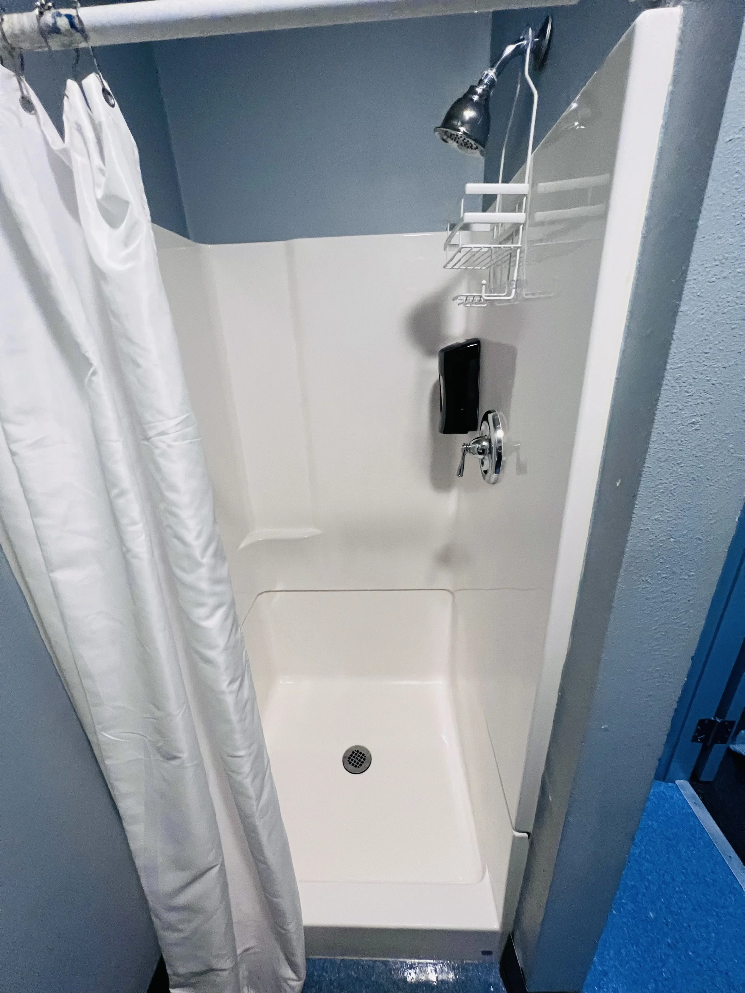Small white shower stall with a curtain on the left, a handheld showerhead, a soap dispenser, and a wire shelf, with blue painted walls.
