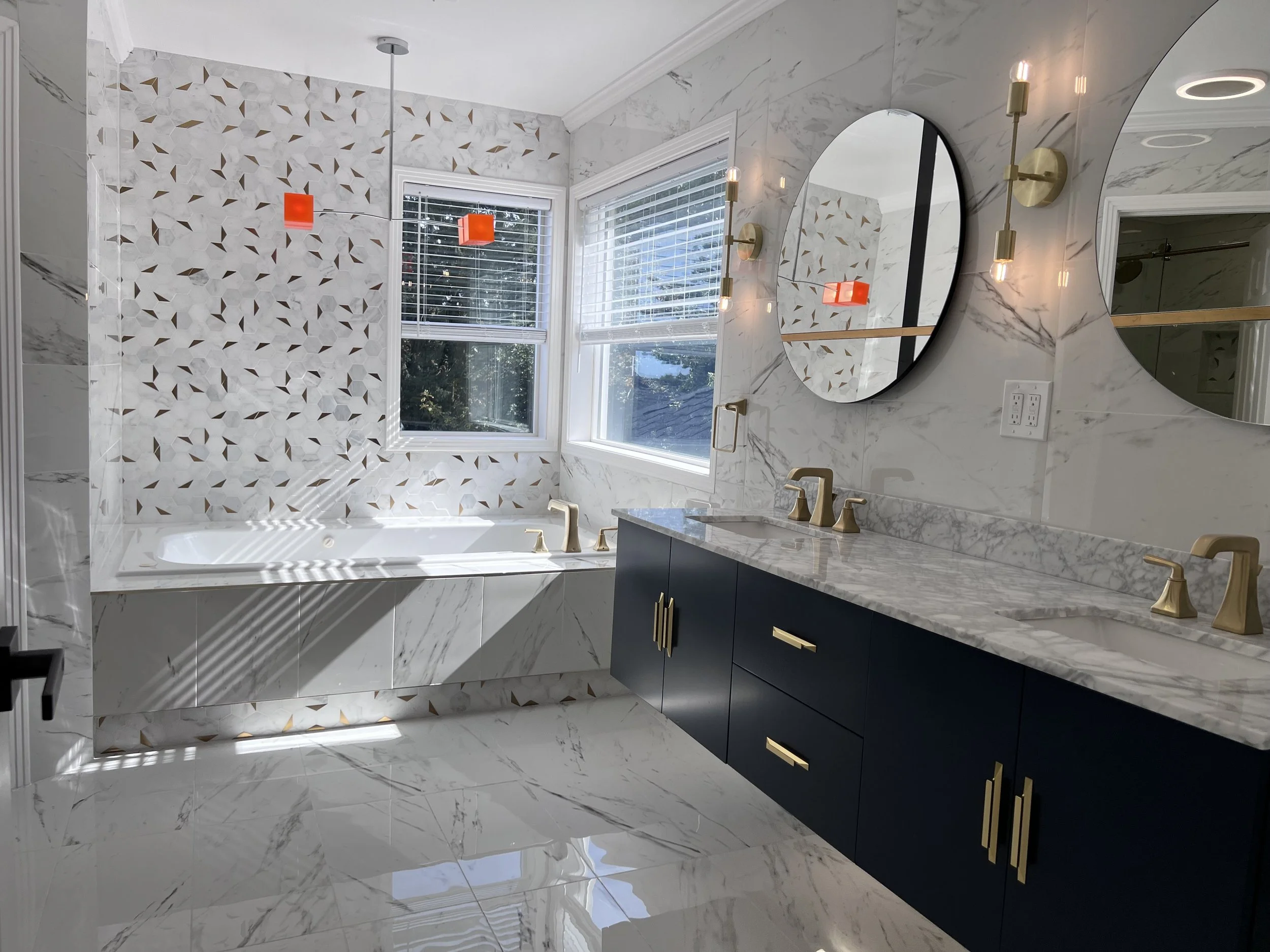 Modern bathroom with marble floors and walls, a large built-in bathtub with a decorative tile wall behind it, two windows with blinds, two round mirrors above a marble countertop with two gold faucets, and gold light fixtures on the wall.