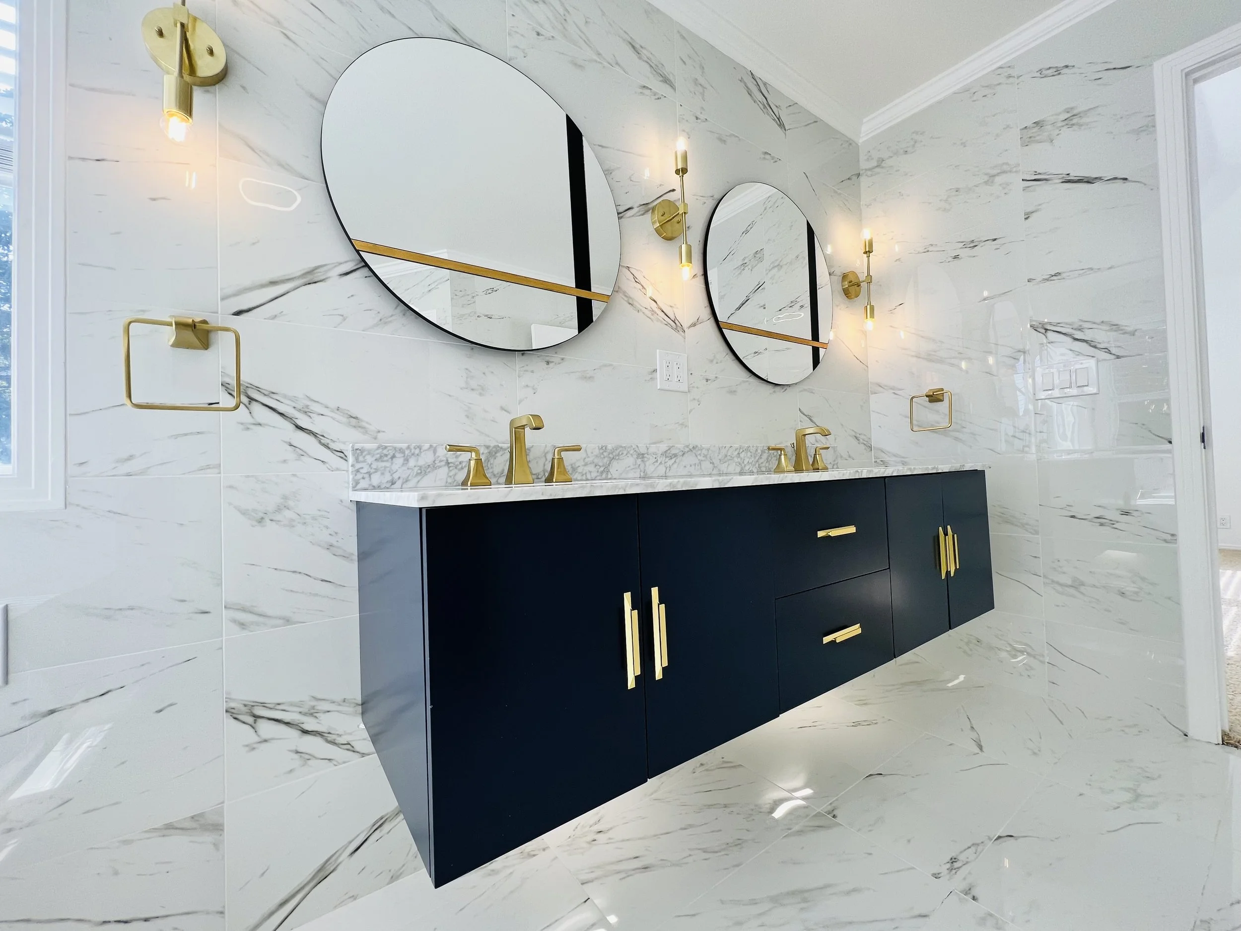 Modern bathroom with marble walls and floor, a dark blue floating vanity with gold handles, two round mirrors, gold fixtures, and wall-mounted light fixtures.