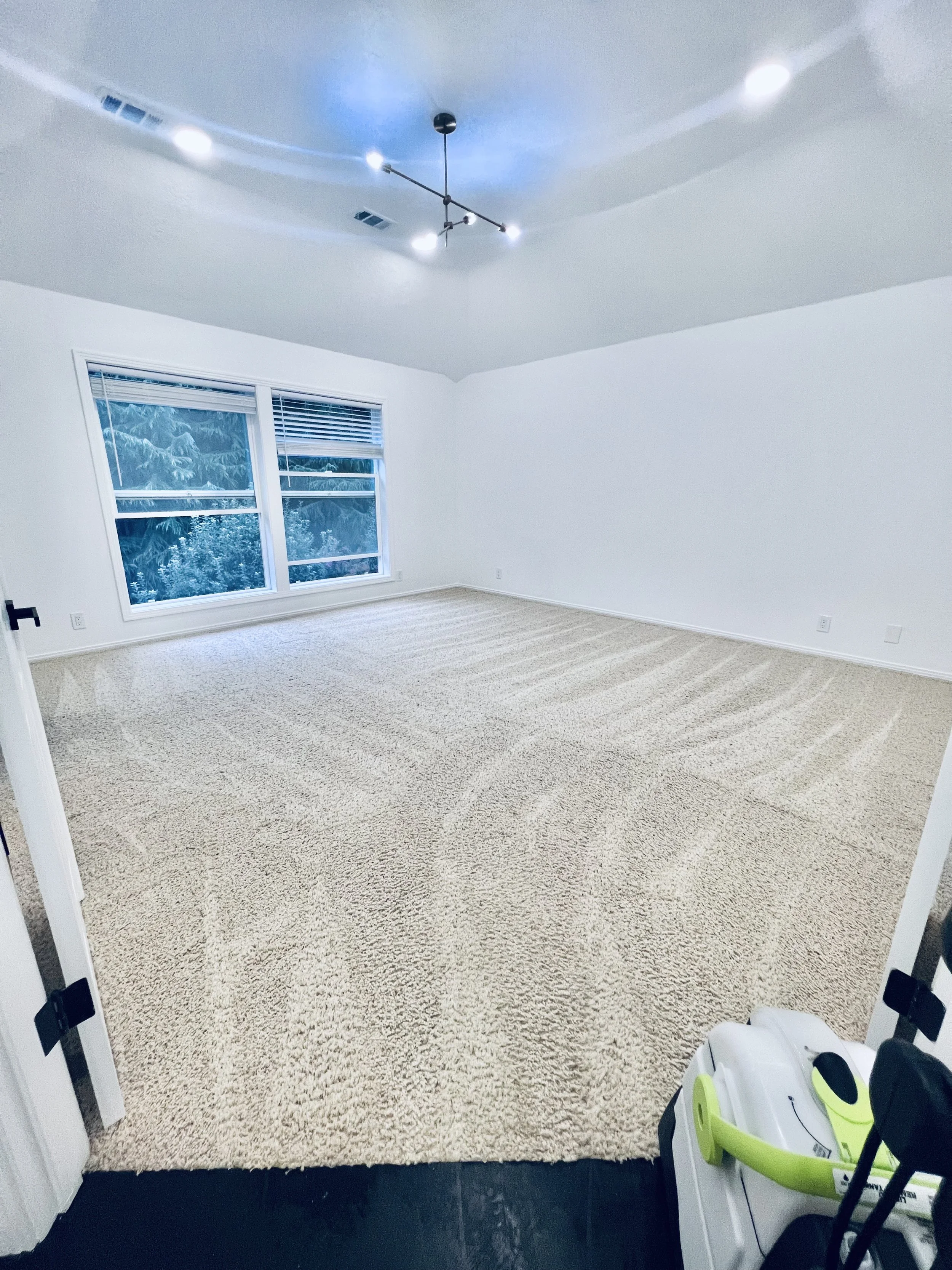 Empty room with beige carpet, white walls, large window, ceiling light fixture, and a small green and white vacuum cleaner near the door.