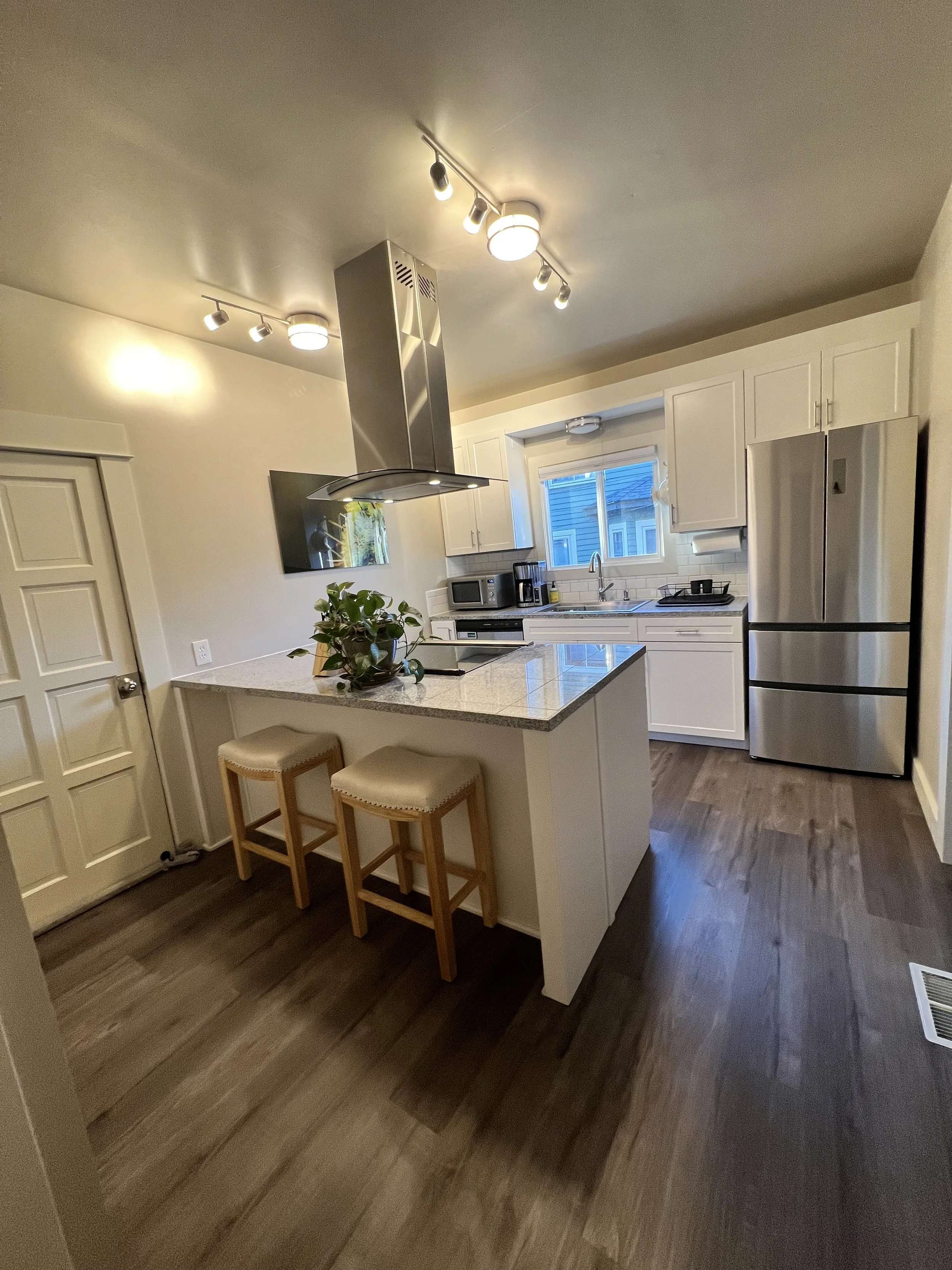 Modern kitchen with white cabinets, stainless steel appliances, a granite island with two stools, and wood flooring.