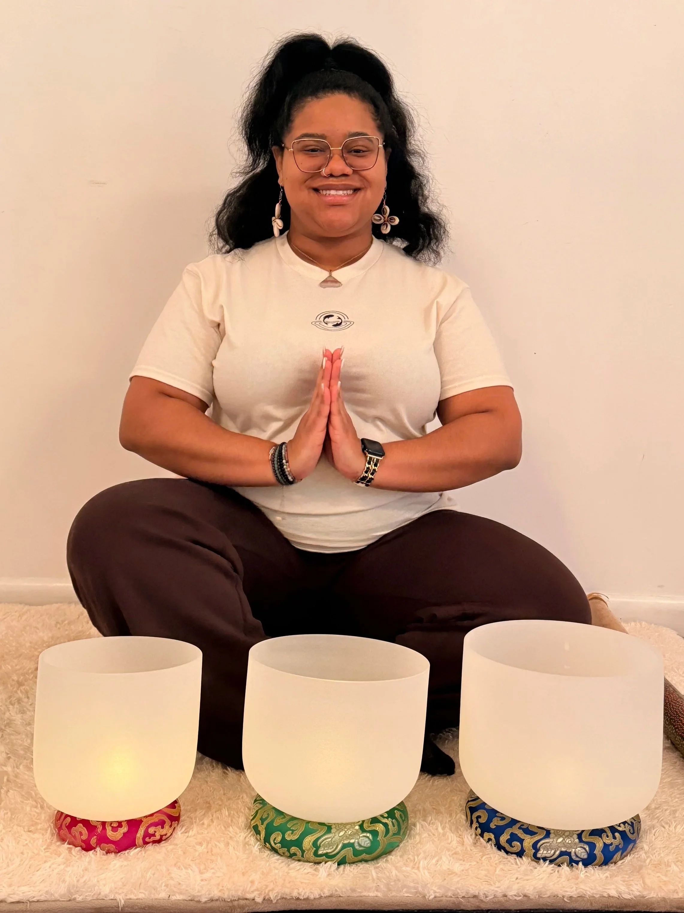 woman sitting on a cushion behind three crystal singing bowlsw and with her hands in prayer position.