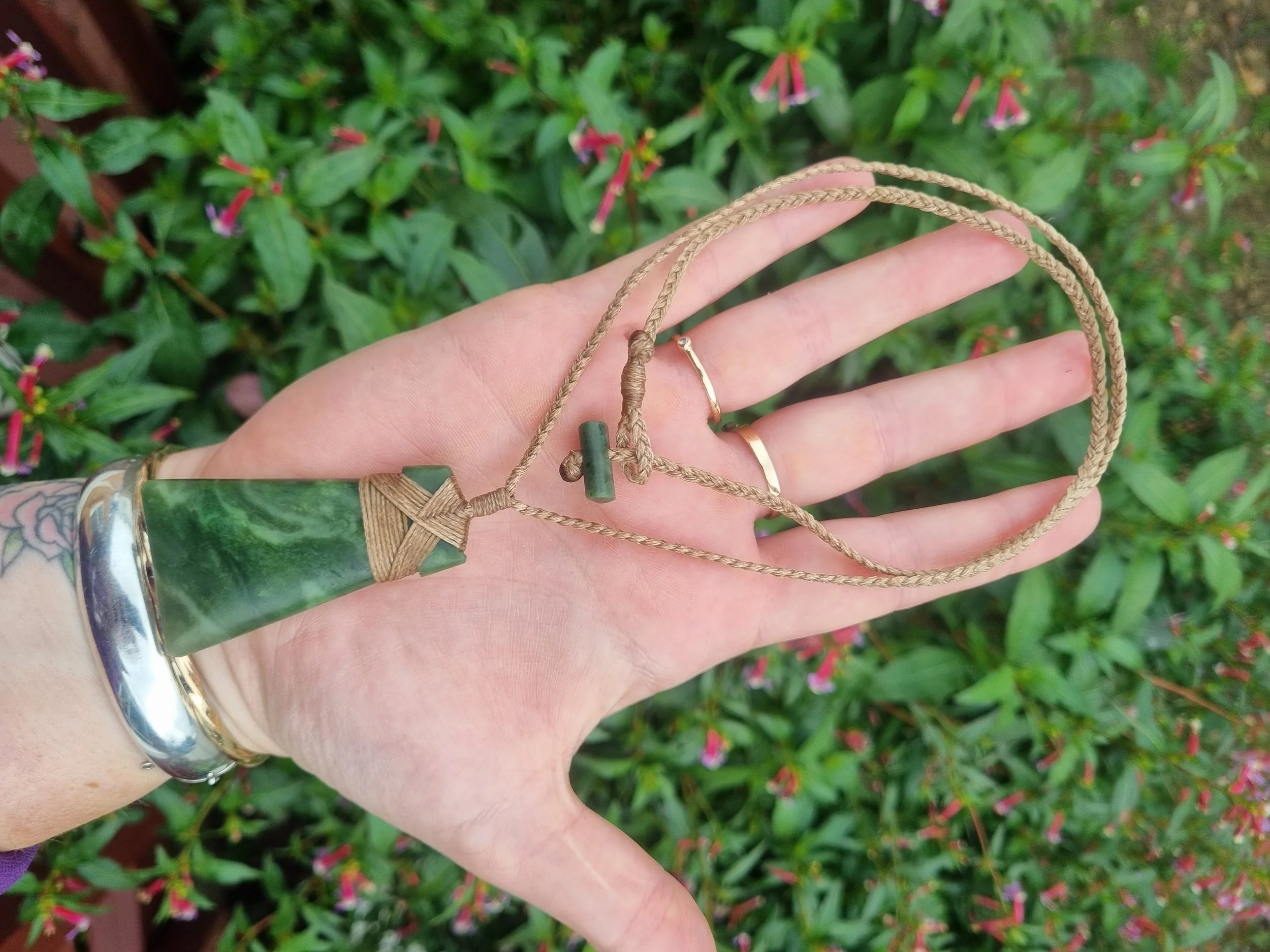Hand holding green gemstone pendant necklace with brown cord, with small rings on fingers, in front of green plants with pink flowers.