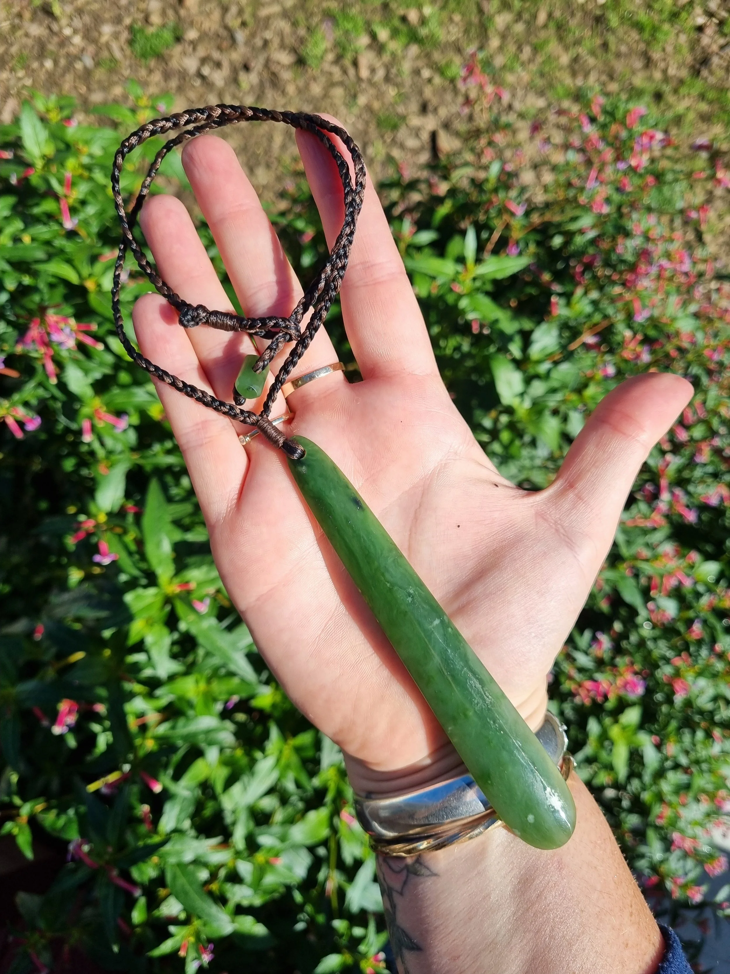 Hand holding a green jade pendant on a black cord necklace with a background of green foliage and pink flowers.