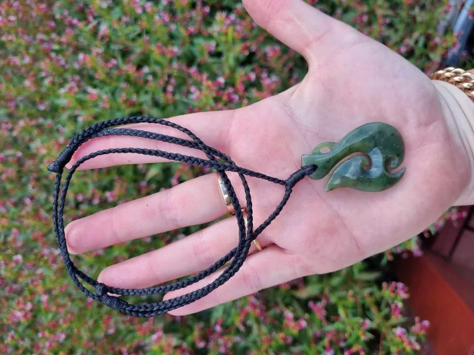 A person holding a black braided necklace with a green jade pendant shaped like a stylized leaf or wave, with pink flowers and green bushes in the background.