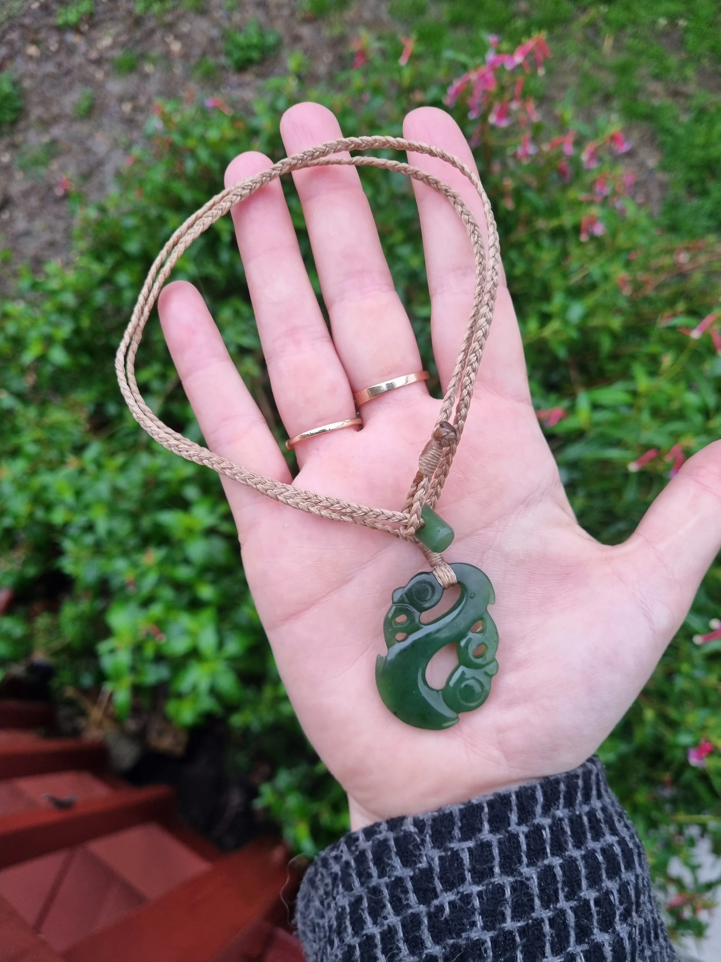 A hand holding a jade necklace with a carved pendant, against a garden background with green plants and pink flowers.