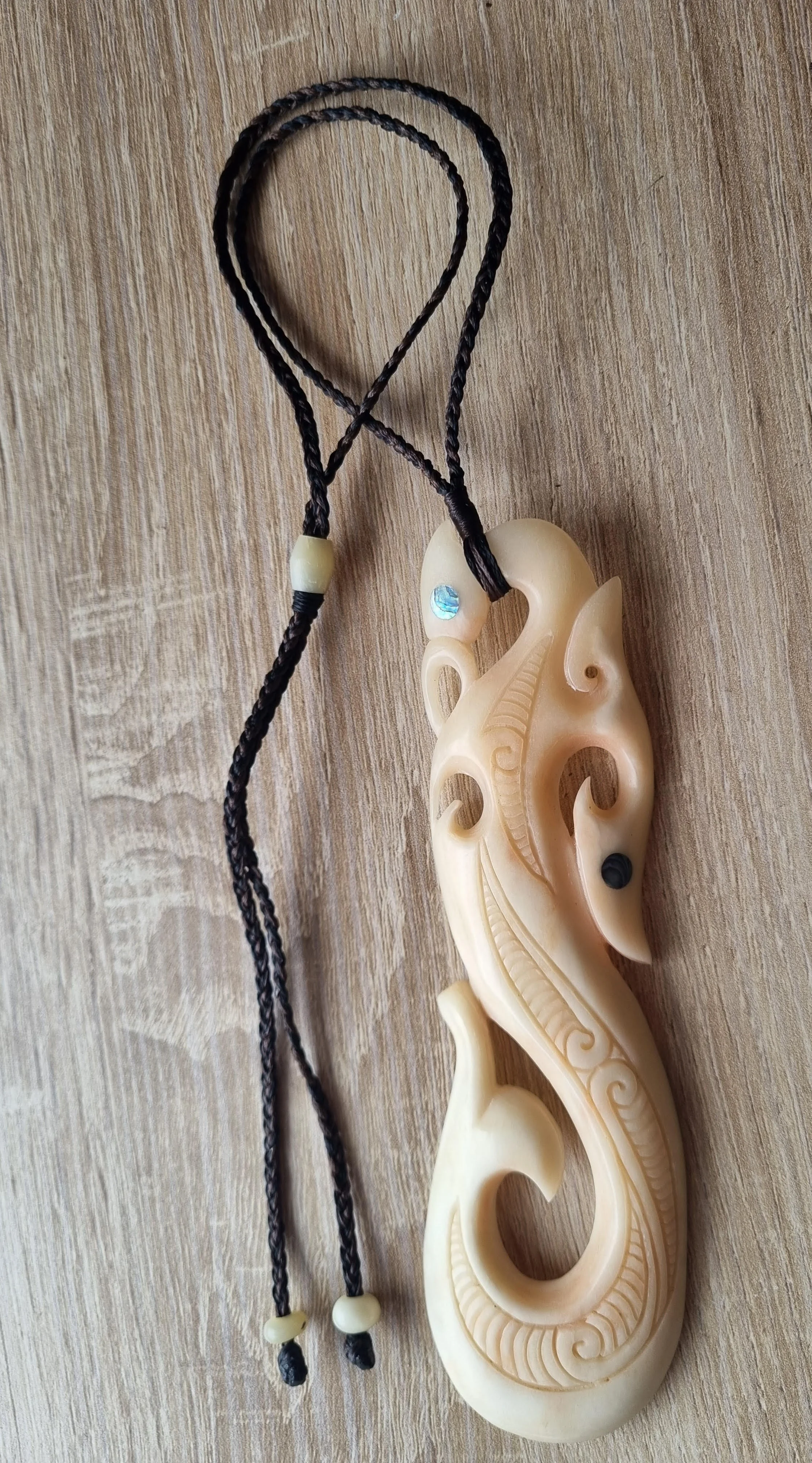 A carved ivory pendant shaped like a fish with intricate patterns, attached to a black braided cord with small beads, laid on a wooden surface.