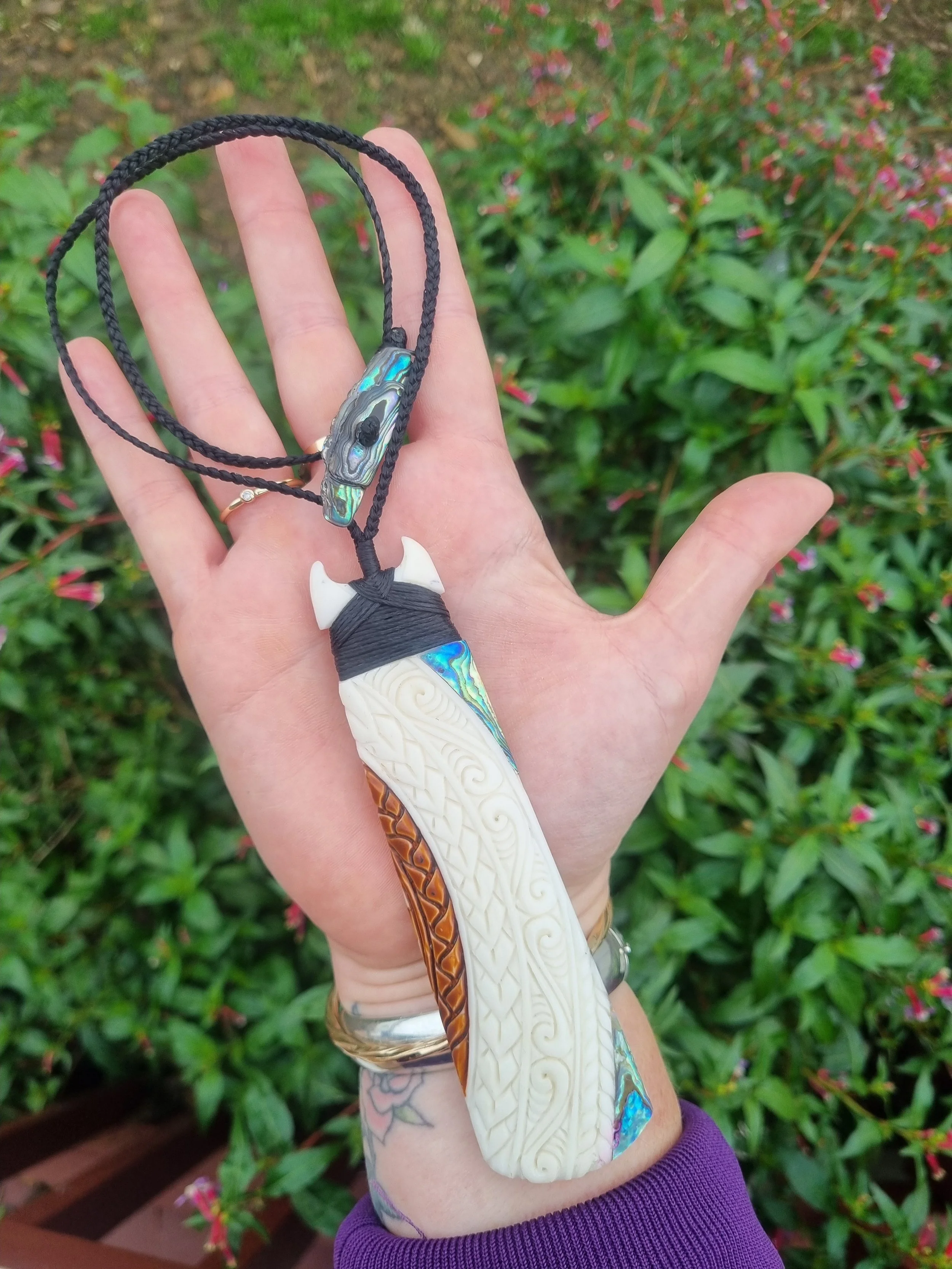A hand holding a Maori-style carved and beaded pendant necklace with a black cord, featuring an intricately carved ivory-colored piece with a smaller iridescent bead on top, against a background of green foliage with pink flowers.