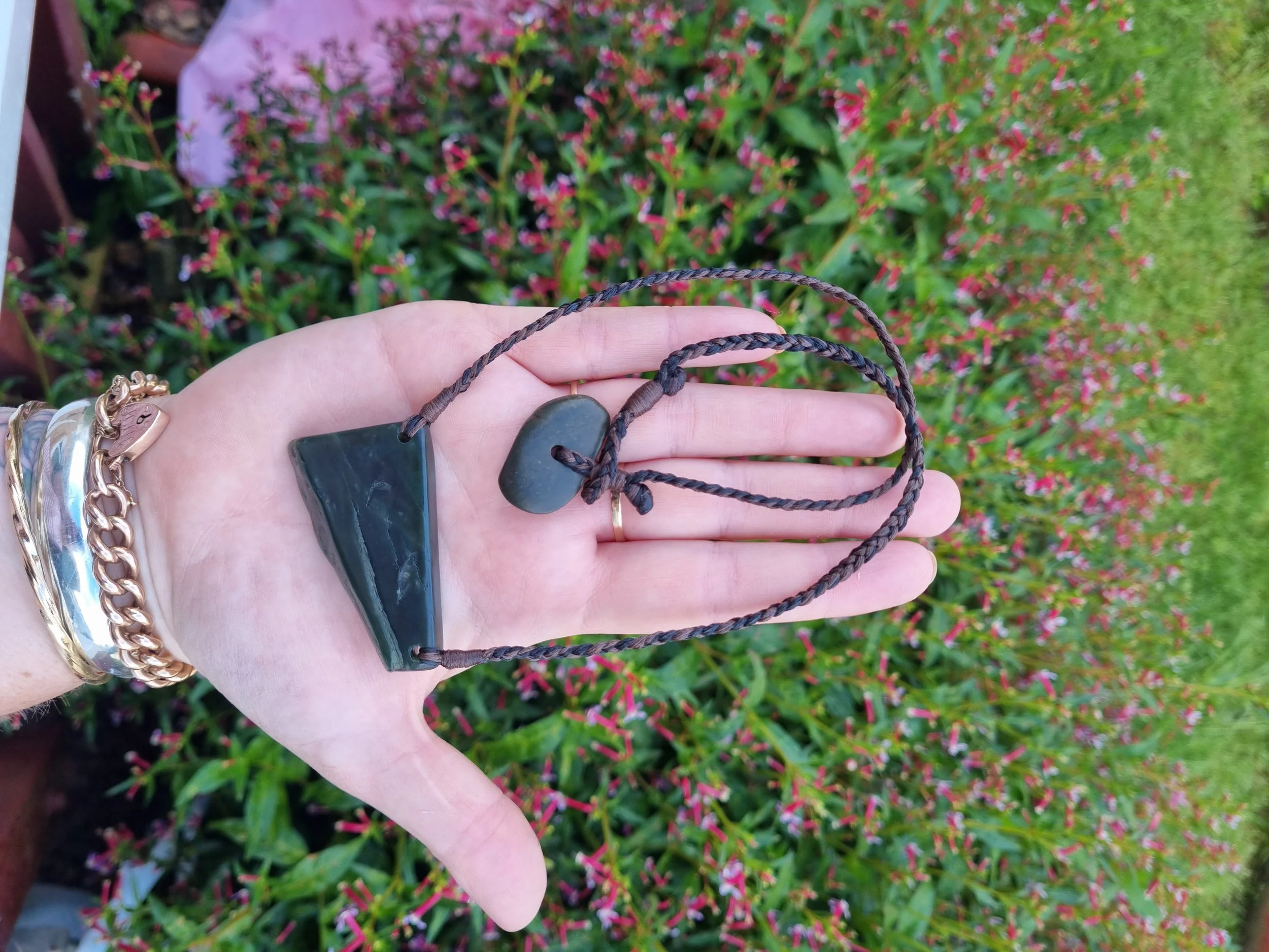 A hand holding a necklace with two black stones, against a background of pink and green flowering plants.