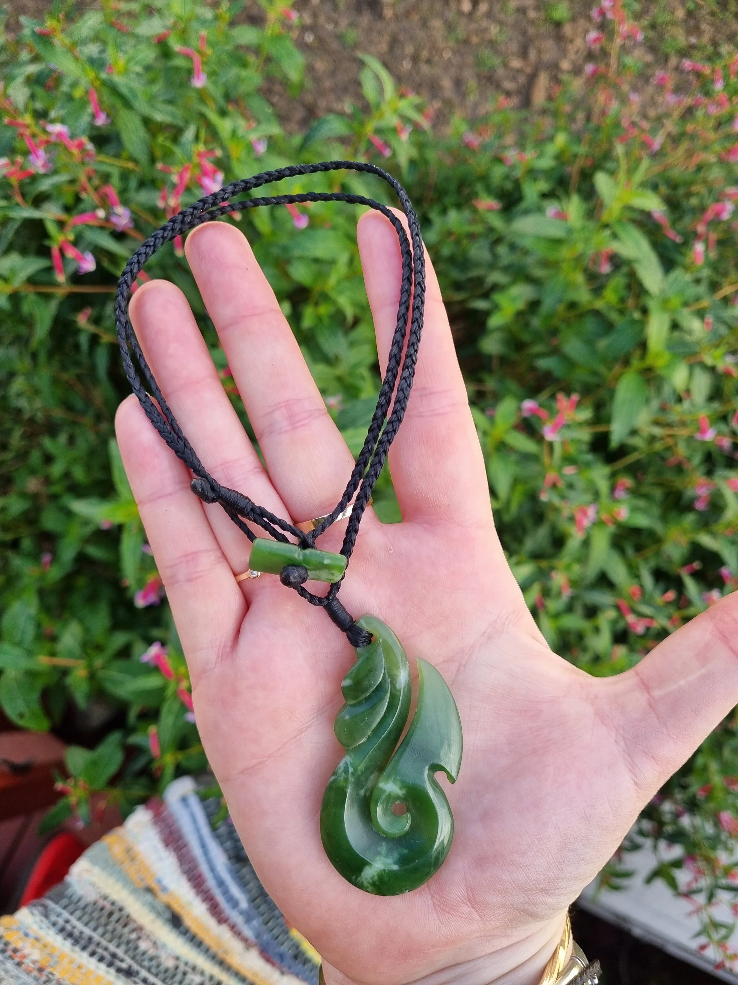 Hand holding a green jade necklace with a carved pendant over a background of green plants and pink flowers.