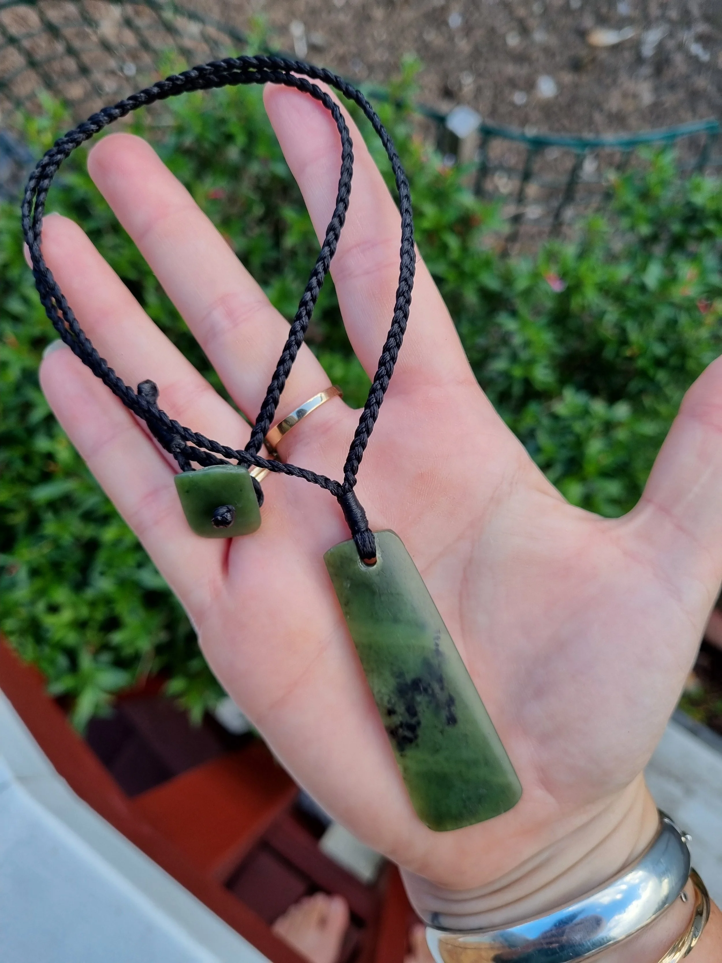 A person holding two jade necklaces with rectangular pendants, one larger and one smaller, against a background of green shrubs and a garden.