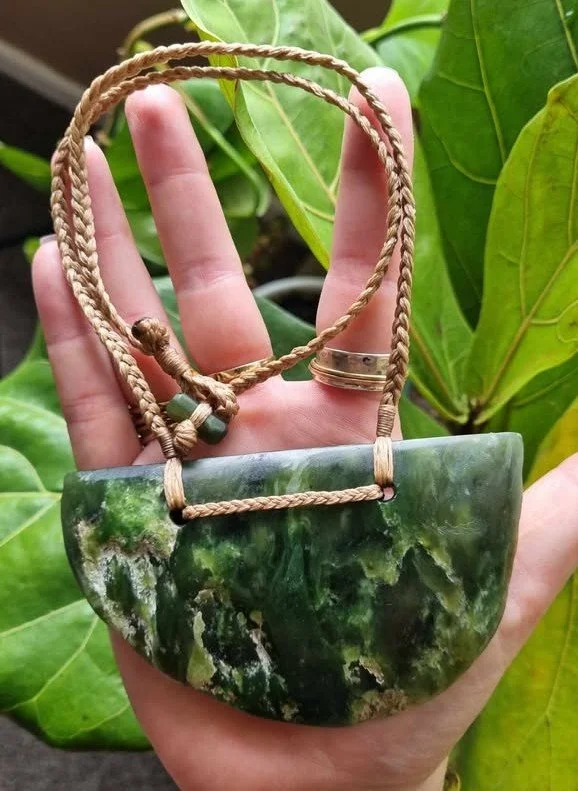 A person holding a green and black marbled pendant with a tan braided cord, set against a leafy background.