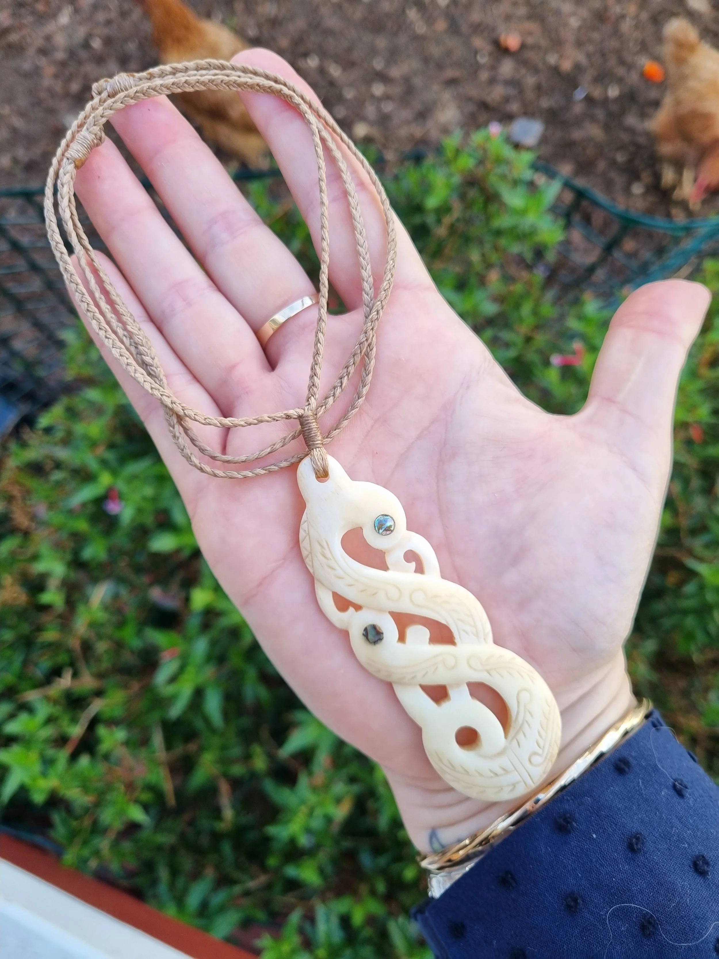 A hand holding a carved ivory or bone seahorse-shaped pendant on a tan cord necklace with small dark stones embedded in the pendant. The background shows dirt and some chickens.