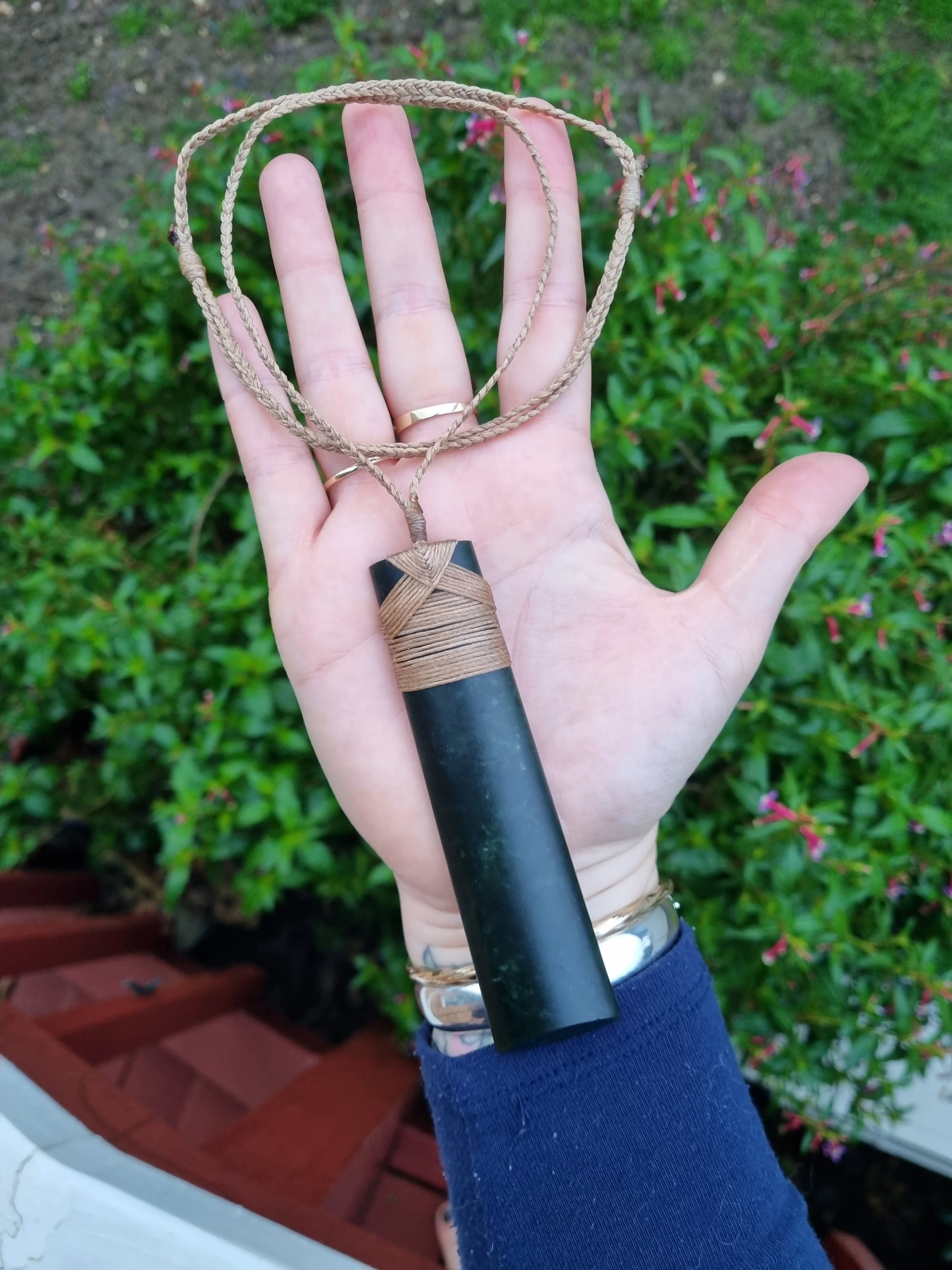 A hand holding a necklace with a large black cylindrical pendant, attached to a braided tan cord, with a background of green plants and pink flowers.