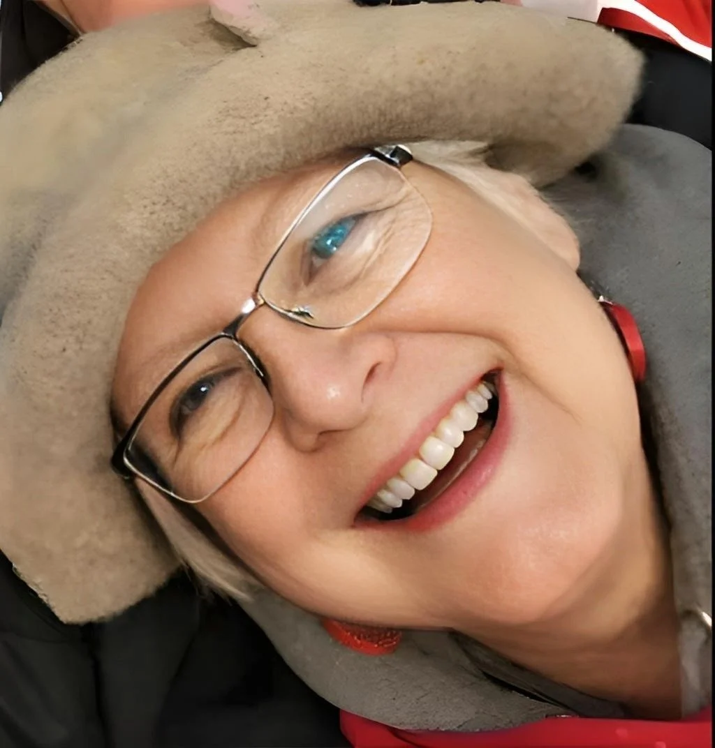 Close-up of a smiling woman with glasses, wearing a beige hat, lying down on a pillow with a gray background.