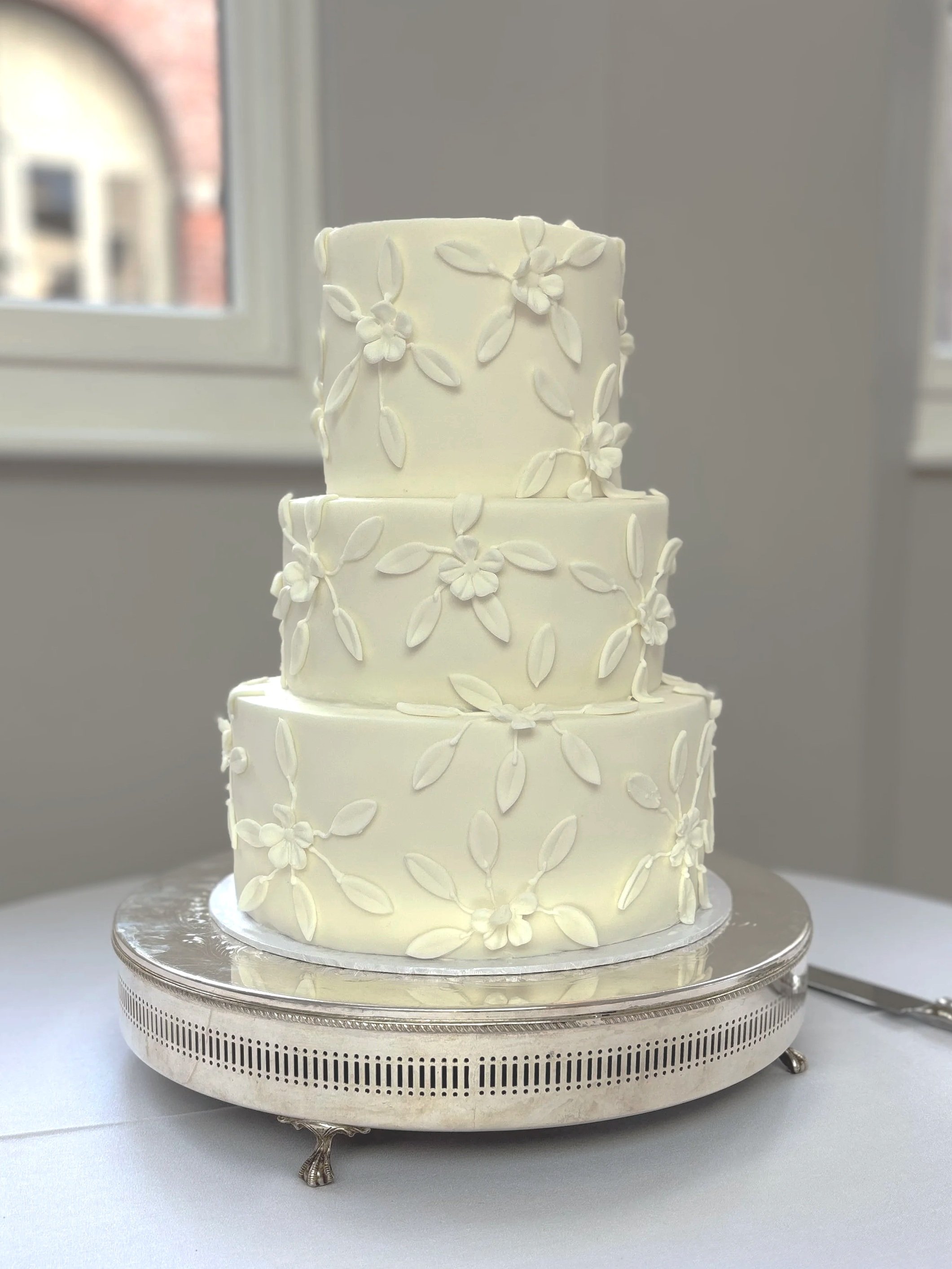 A three-tier white wedding cake decorated with white floral and leaf patterns, placed on a silver cake stand.