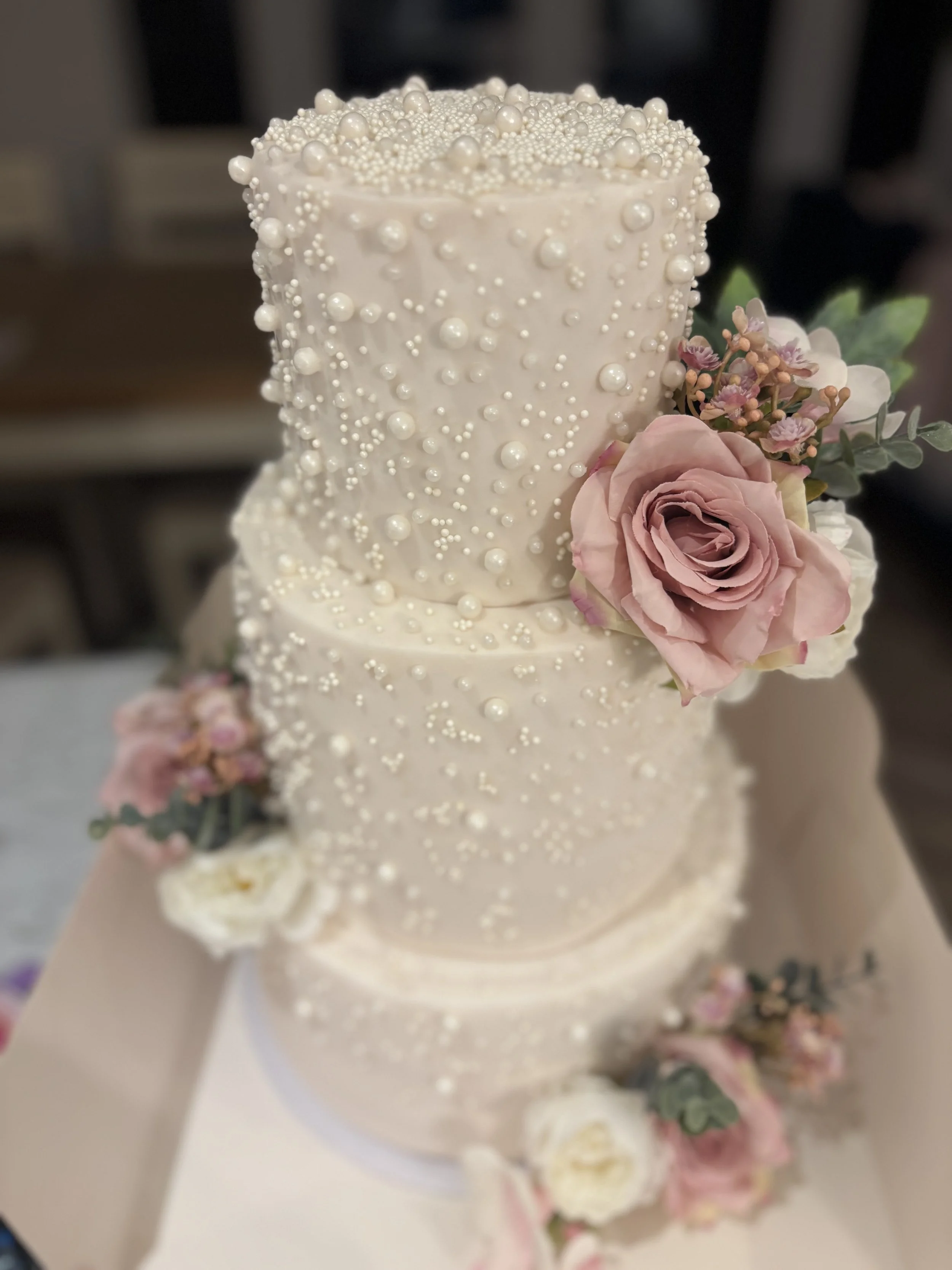 A tall, white wedding cake decorated with numerous small white sugar pearls and pearl-like beads, with fresh pink and white roses and greenery on the side.