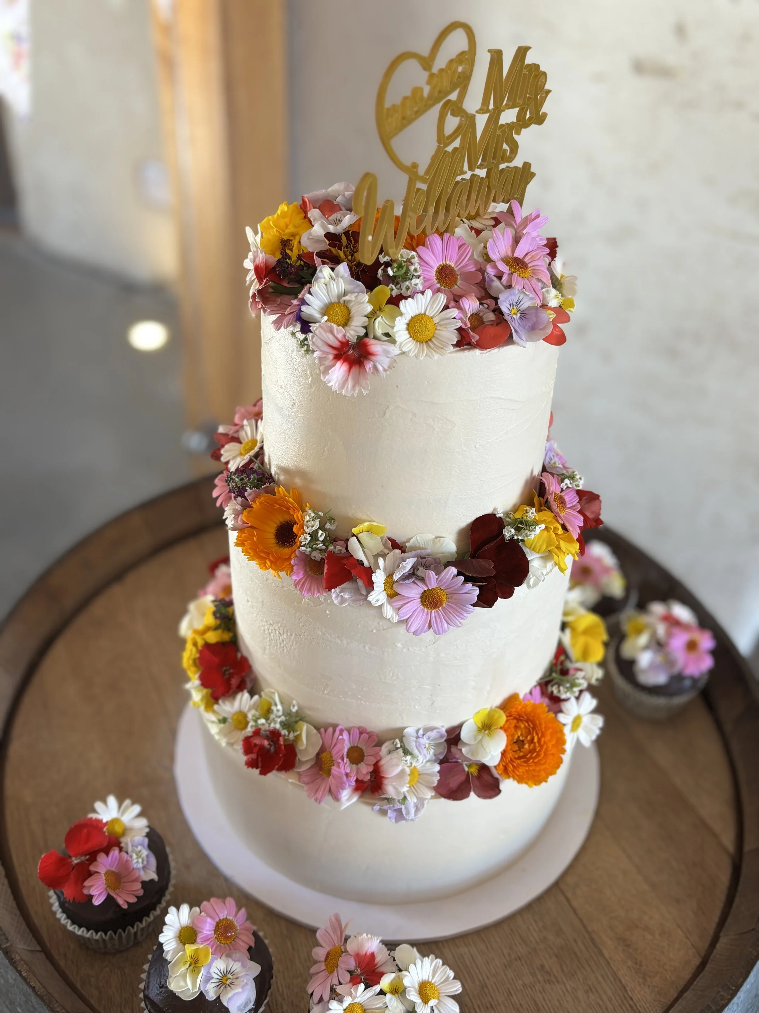 A three-tiered white wedding cake decorated with colorful fresh flowers including daisies, roses, and marigolds, with a gold cake topper that reads 'Mr & Mrs Wellington.' The cake is placed on a wooden table surrounded by cupcakes decorated with similar flowers.