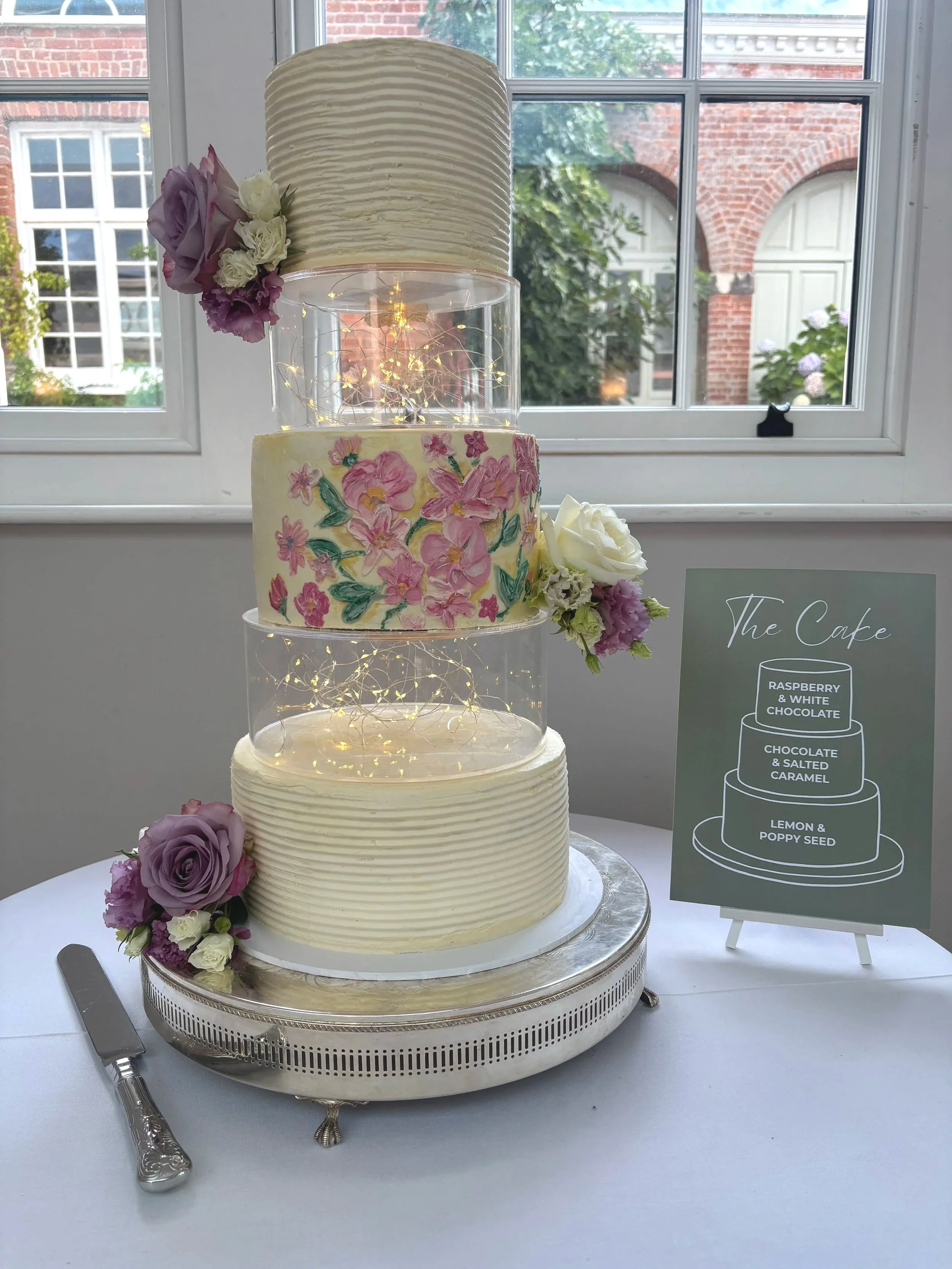 A multi-tiered wedding cake with white ribbed icing, decorated with fresh purple and white flowers, and small lights inside the middle tiers, placed on a silver cake stand next to a silver cake knife, with a sign listing cake flavors including raspberry and white chocolate, chocolate and salted caramel, lemon and poppy seed.