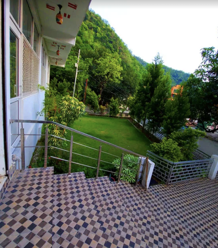 View from a balcony with checkered floor tiles, overlooking a green lawn, trees, and mountains in the distance.