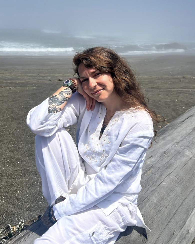 Woman with long curly hair, tattoo on her arm, wearing white clothing, sitting on a wooden log at the beach, with sand and ocean waves in the background.