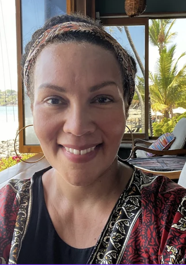 Woman smiling indoors with tropical trees and water visible through open windows behind her.