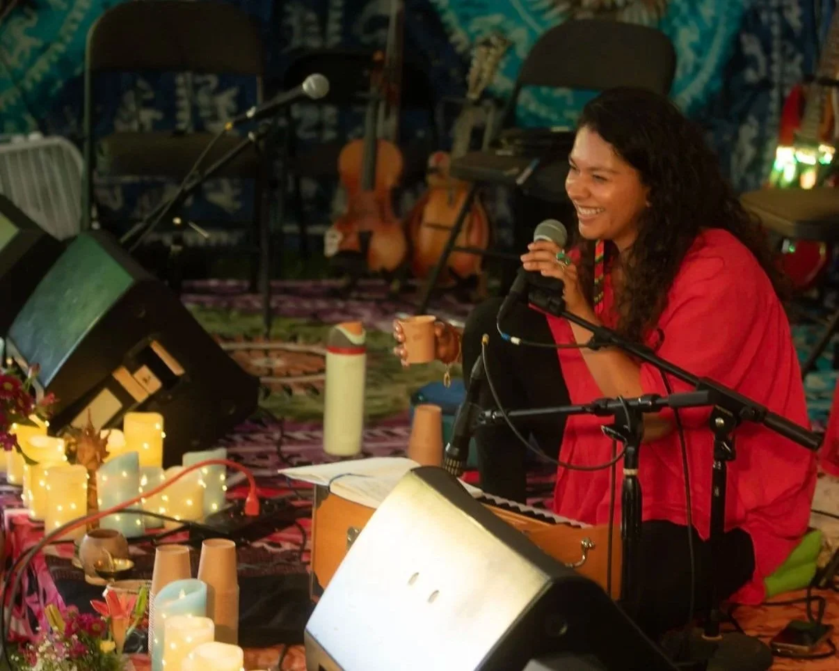 A woman with curly dark hair wearing a red top, sitting on the floor with musical instruments and candles, holding a microphone and smiling during a performance in a cozy, decorated indoor space.