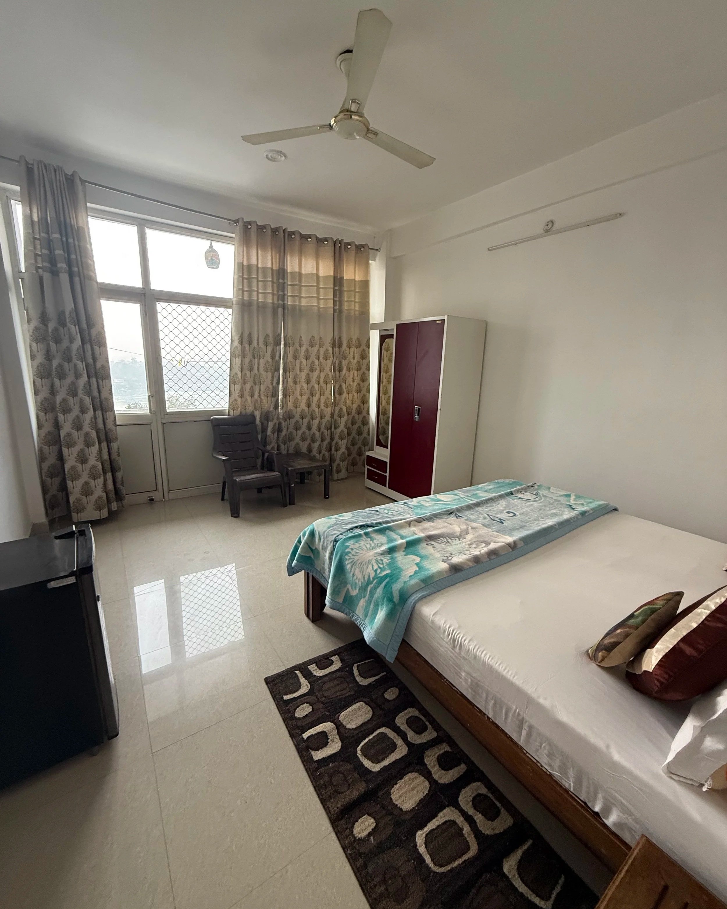 A bedroom with a bed, a patterned rug, a small cabinet, beige curtains, a ceiling fan, a chair, and a window with a view outside.
