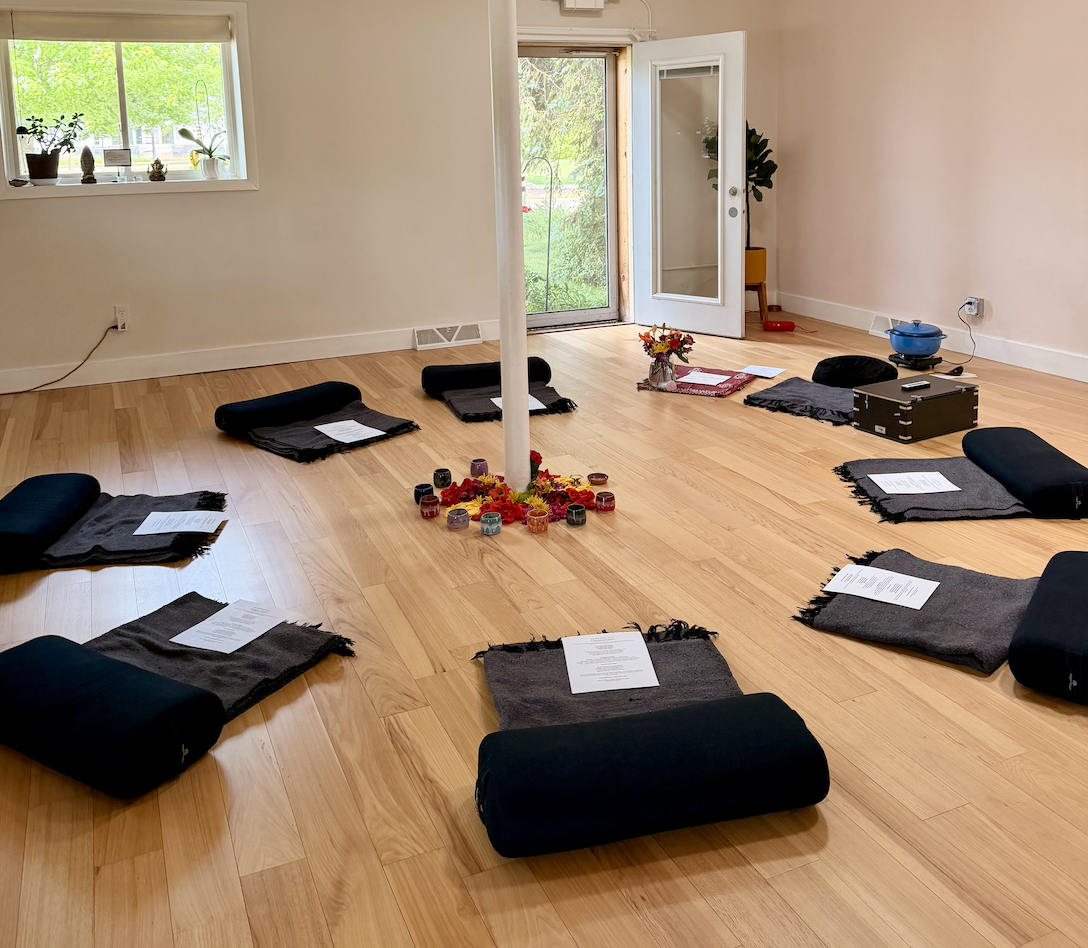 A circular arrangement of cushions with papers on top of each cushion, set up for a meditation or yoga session in a bright room with wooden floors. In the center, there is a pillar surrounded by small candles surrounded by flowers. To the right, there is an open door leading to an outdoor area with green trees, and a window with plants on the sill.