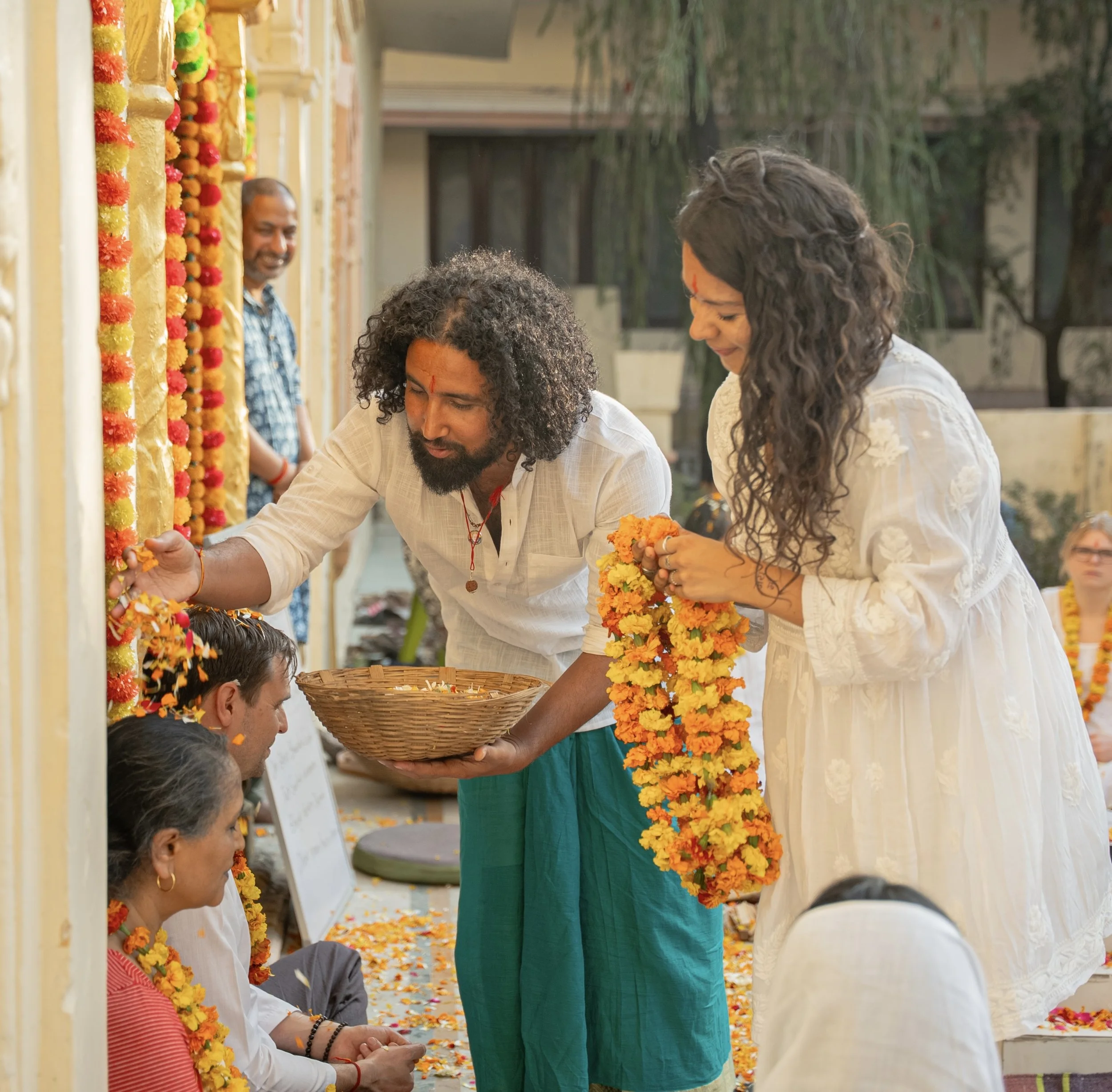 People participating in a traditional Indian ceremonial ritual, with individuals wearing white garments and floral garlands, exchanging offerings, in an outdoor setting decorated with flower garlands.