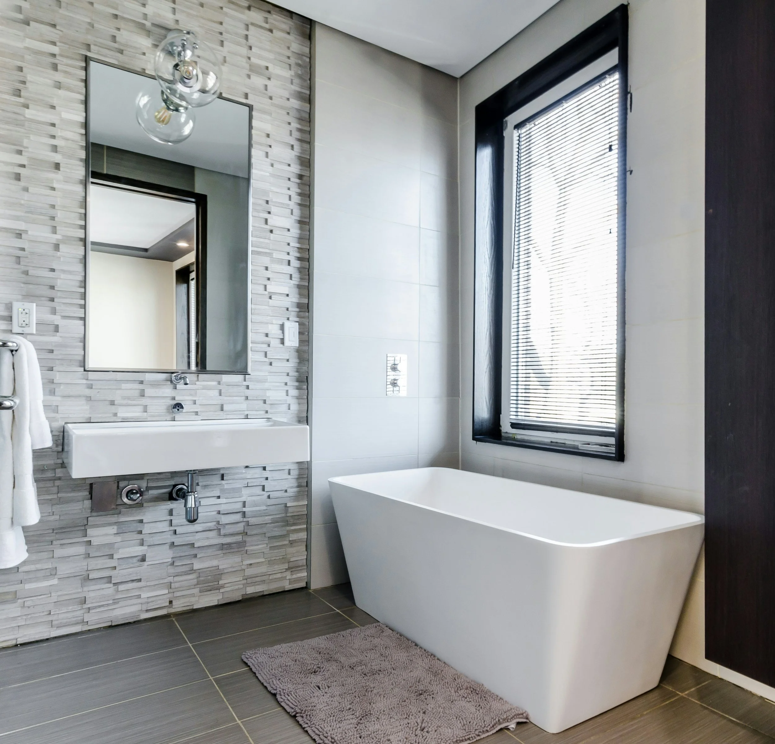 Modern bathroom with a freestanding bathtub, wall-mounted sink, large mirror, textured tile wall, and a window with blinds.