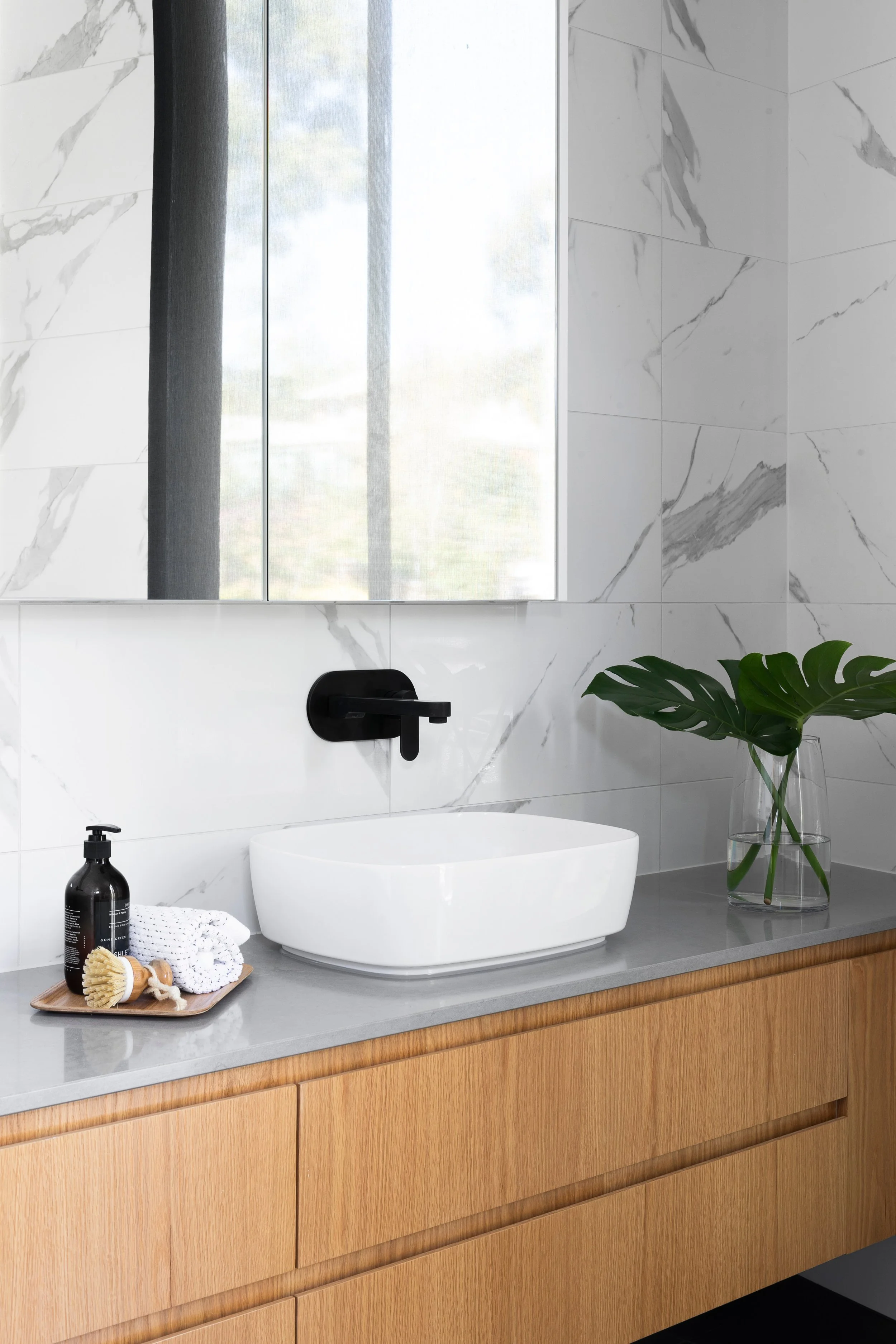 Modern bathroom with a white vessel sink, black wall-mounted faucet, and large mirror. The countertop is gray with wooden cabinets below. Decor includes a tray with a black bottle, white towels, and a wooden brush, as well as a glass vase with green 