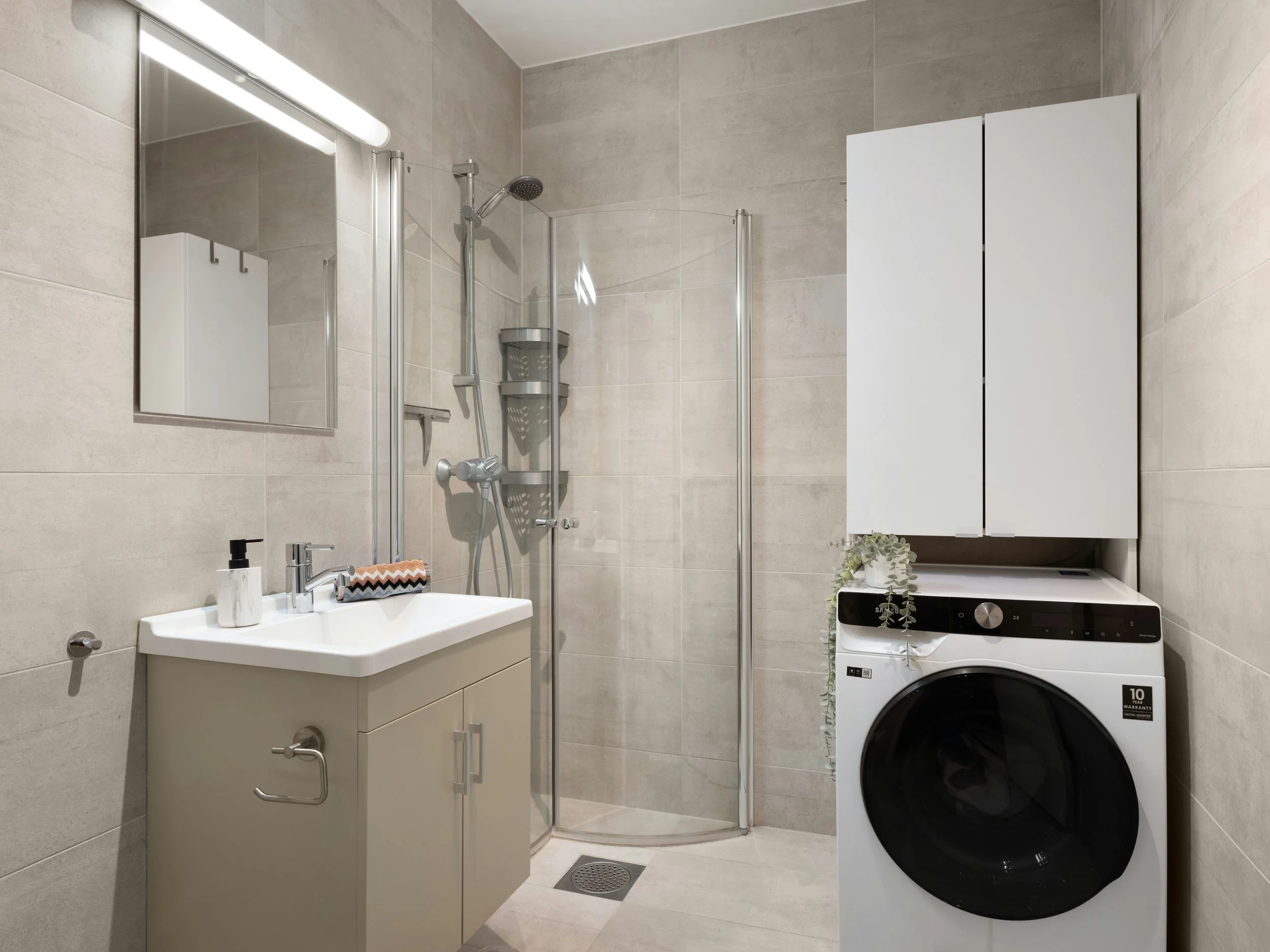 Modern bathroom with a white vanity, wall-mounted mirror, and sink with chrome fixtures. A glass shower enclosure is in the corner. A front-loading washing machine is adjacent to white cabinets. The walls are tiled in a light gray color.