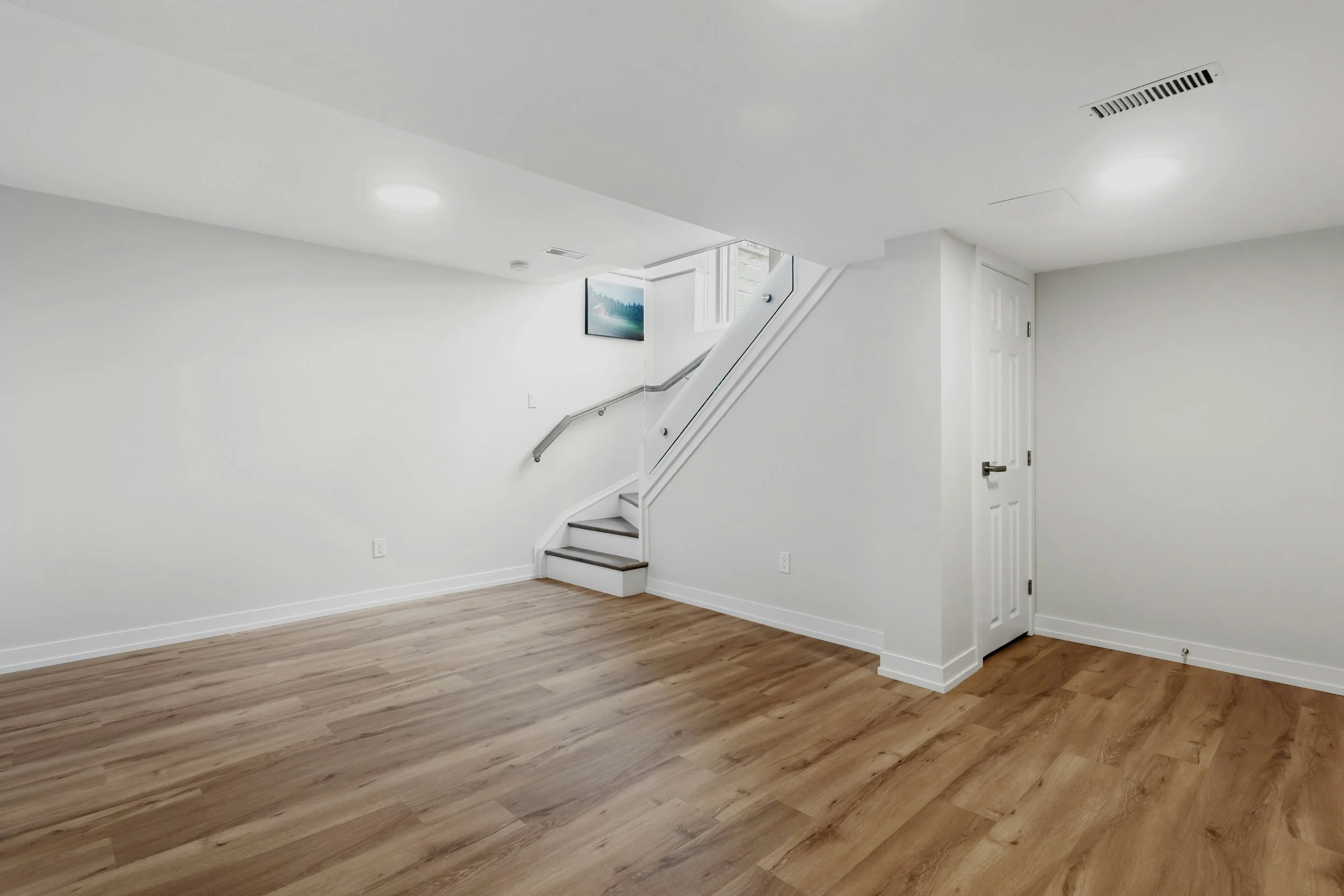 Empty basement with hardwood floor and white walls, featuring staircase and white door.