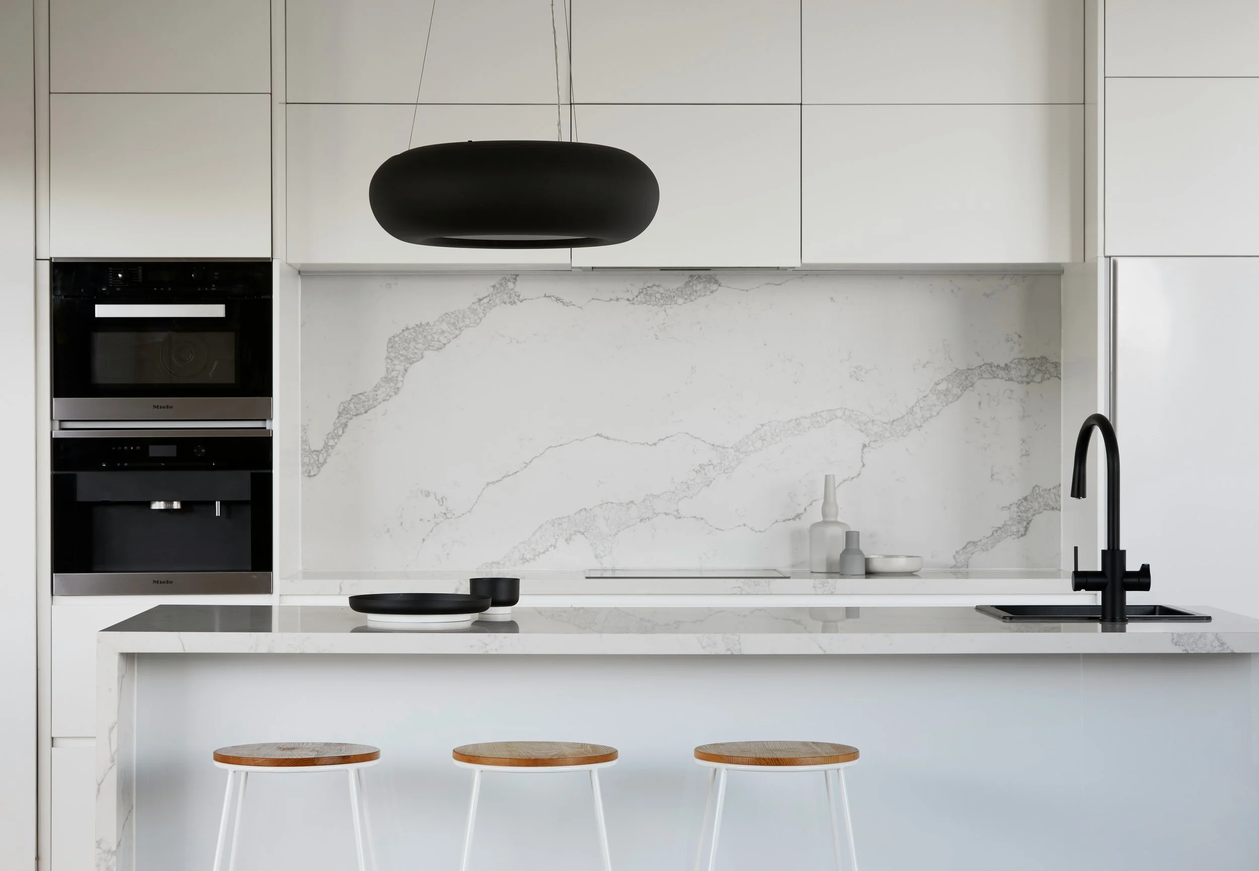 Modern kitchen with white cabinets, marble backsplash, black faucet, three stools, and a hanging light.