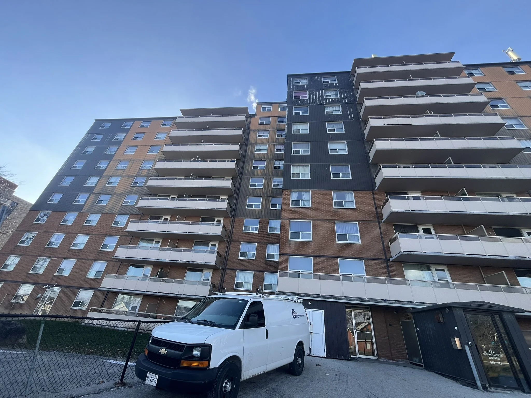 High-rise apartment building with multiple balconies, blue sky, and a white van parked in front.